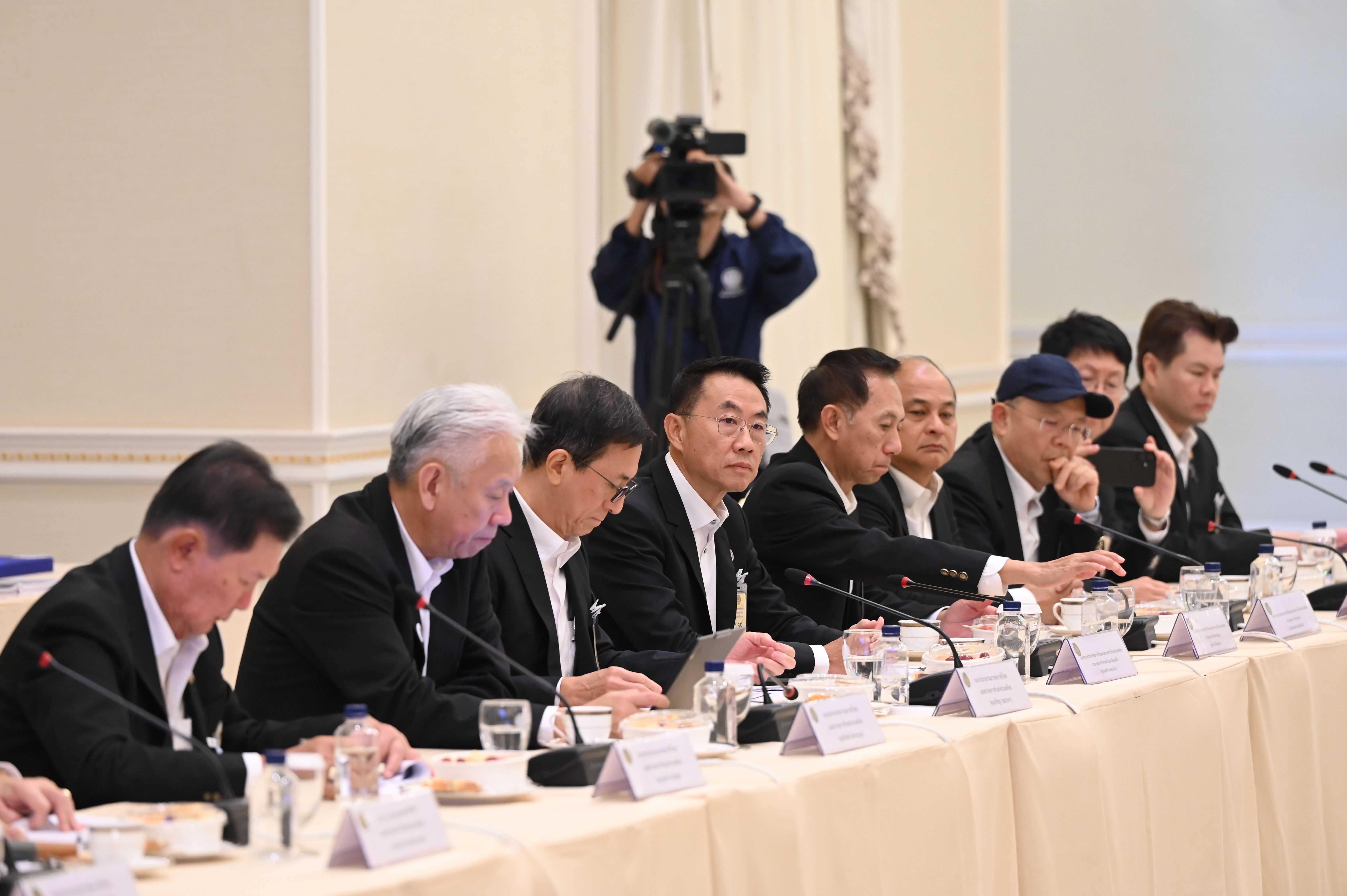 Men in dark suits seated at a long conference table with microphones and a camera.