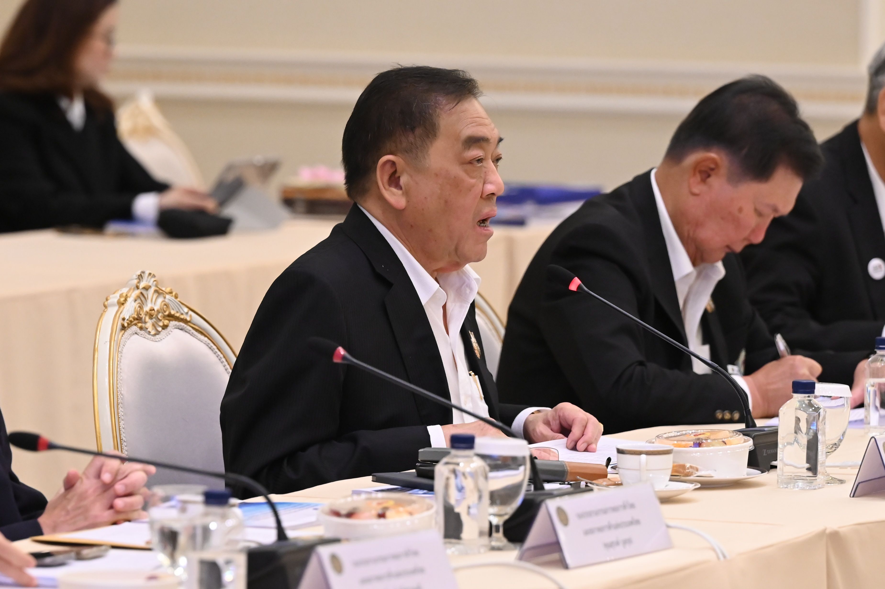 Older Asian man in a dark suit speaking at a formal meeting table.