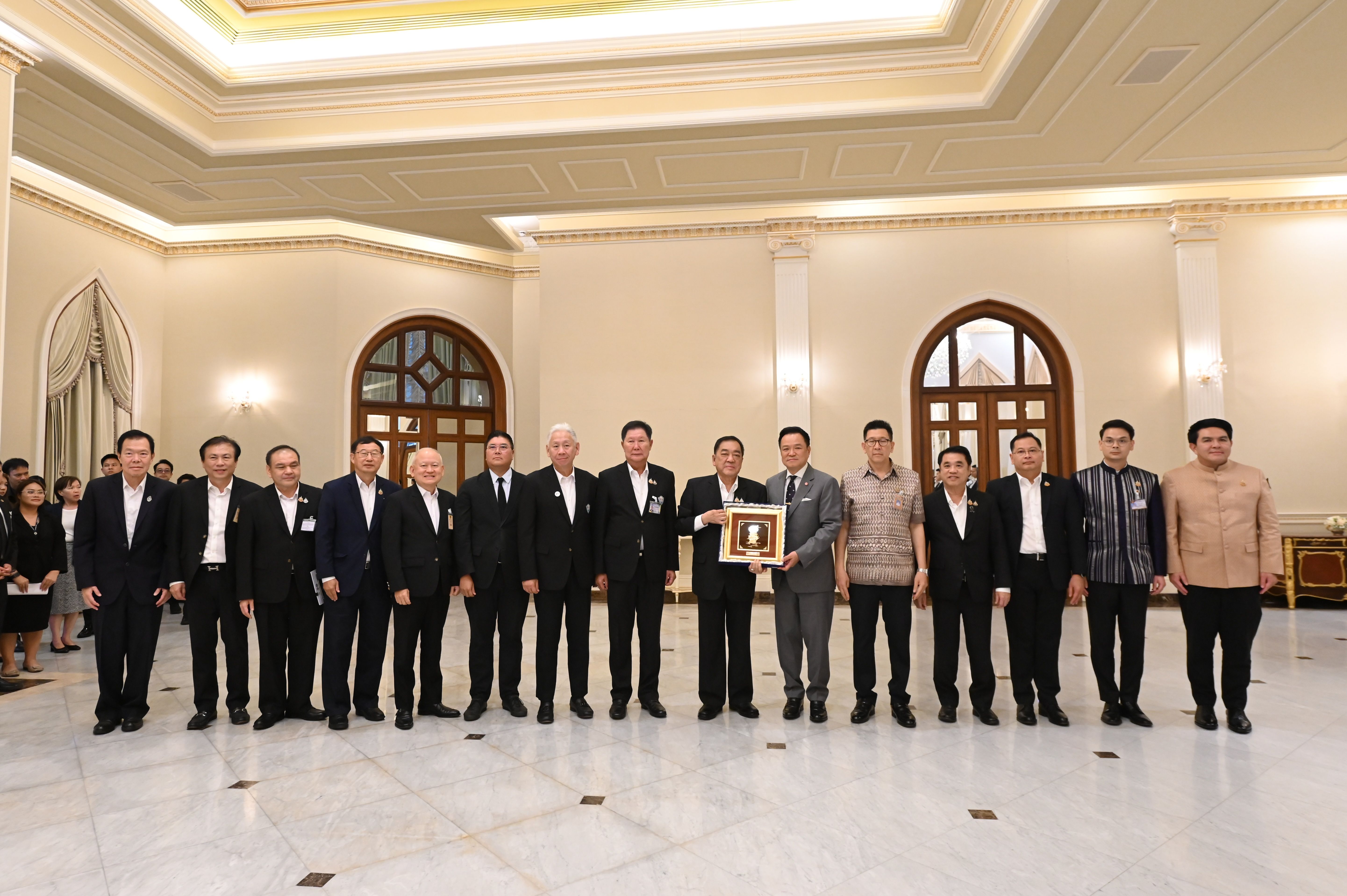 Formal group of men in suits posing with a framed award in an ornate hall.