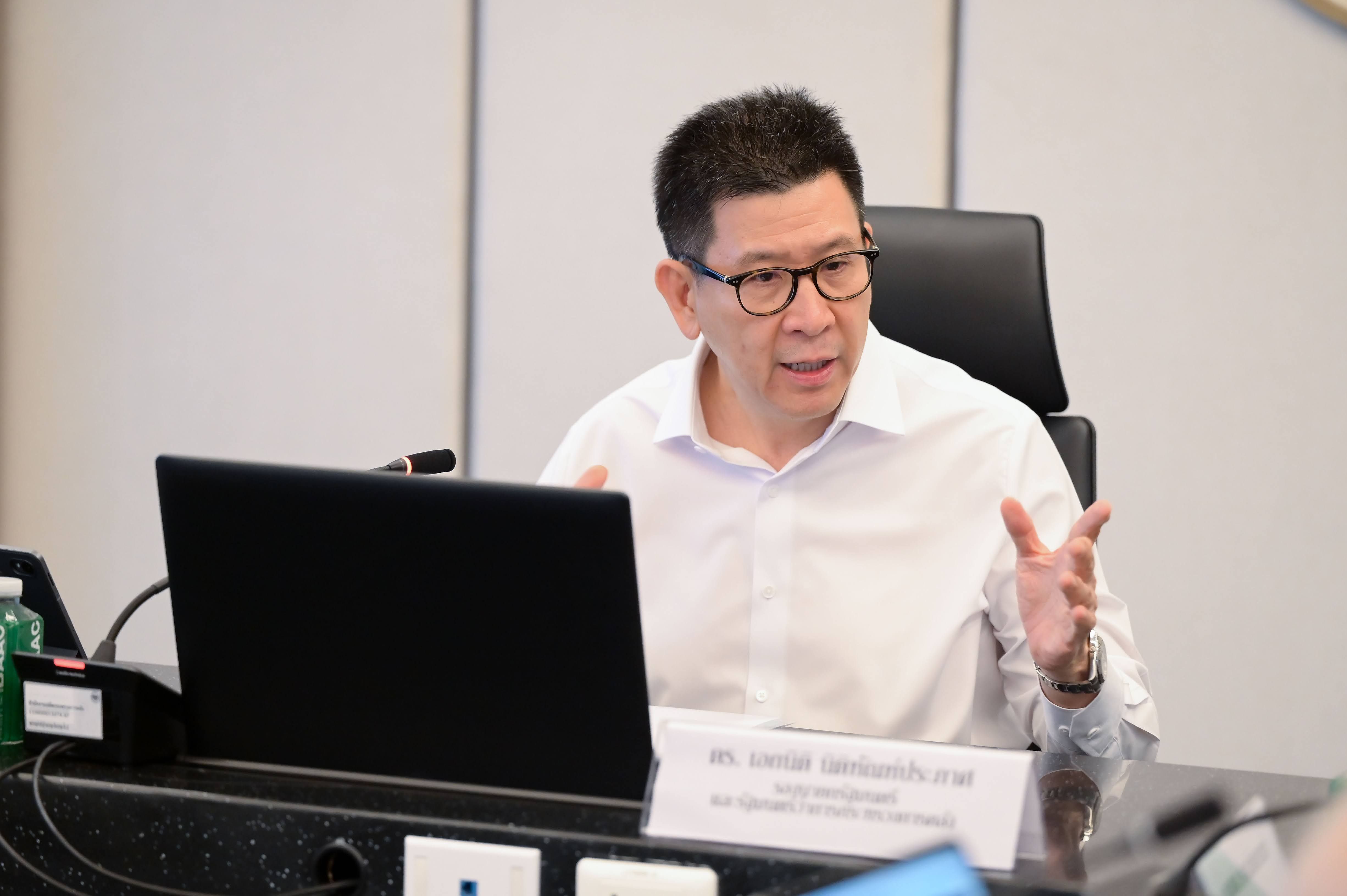 Man in glasses and white shirt gesturing while speaking at a table with a laptop.