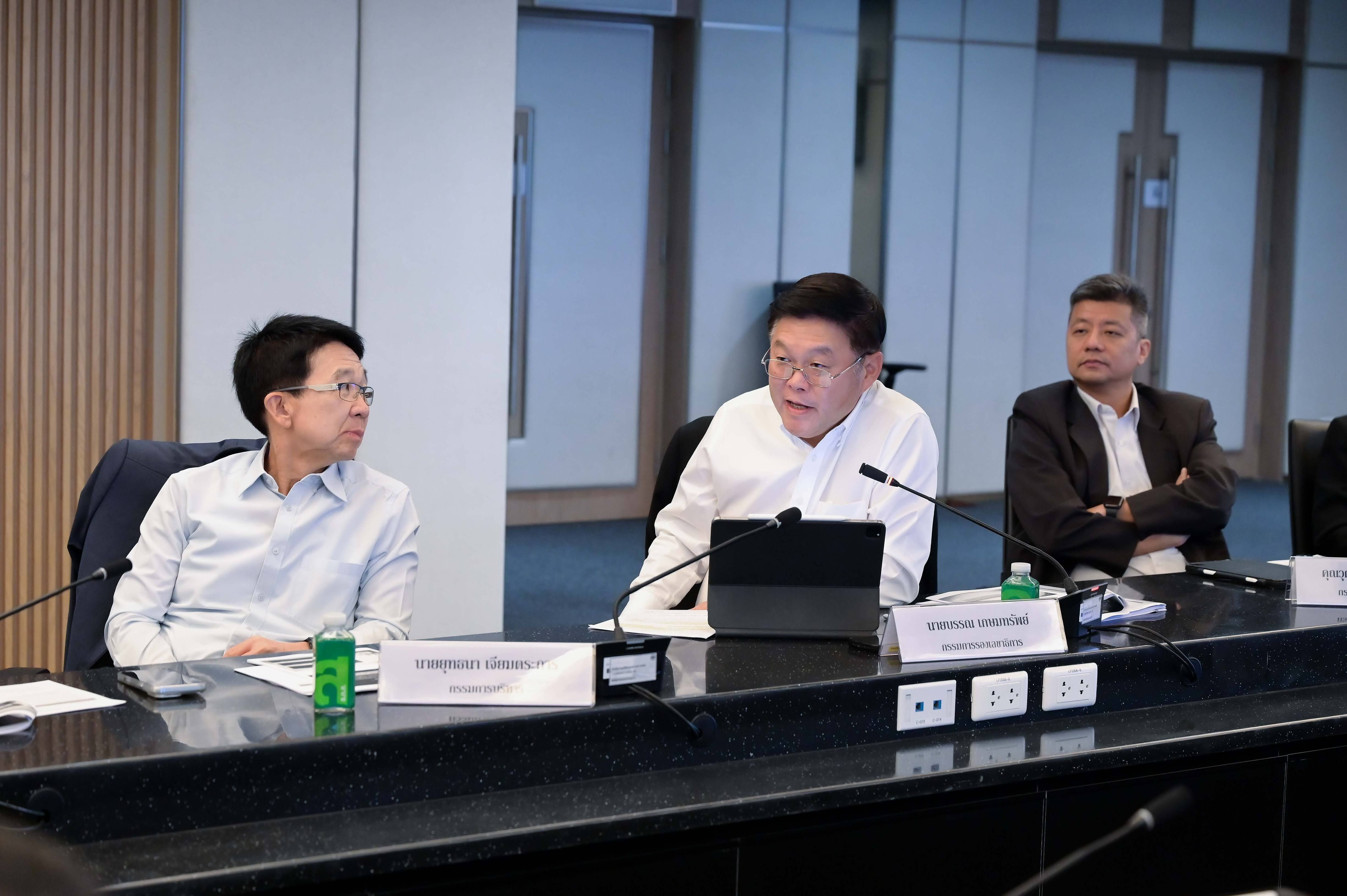 Three men at a conference table, one speaking into a microphone while others listen.