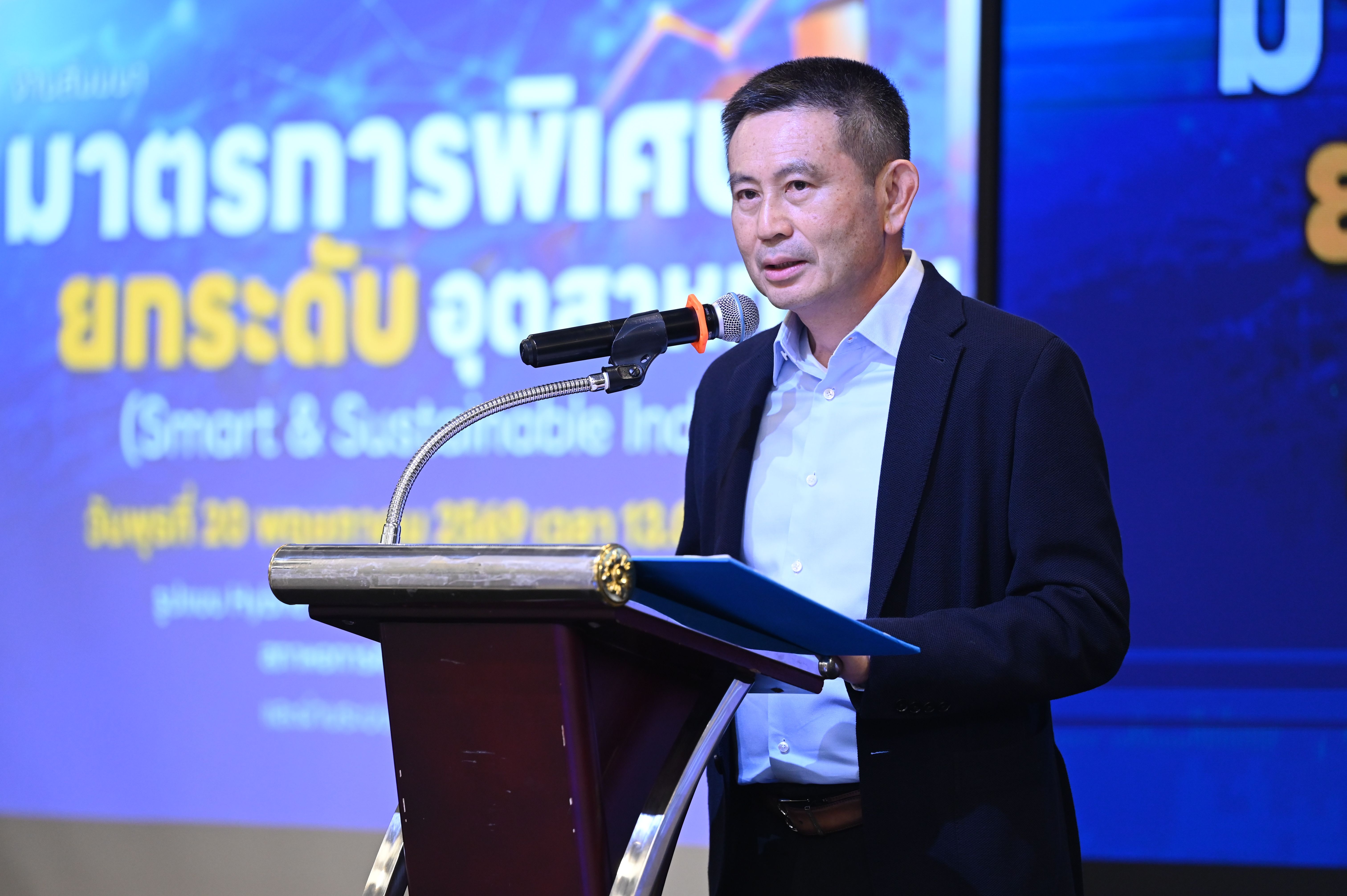 Man in a suit speaks at a podium with microphone, holding a document, blue background.