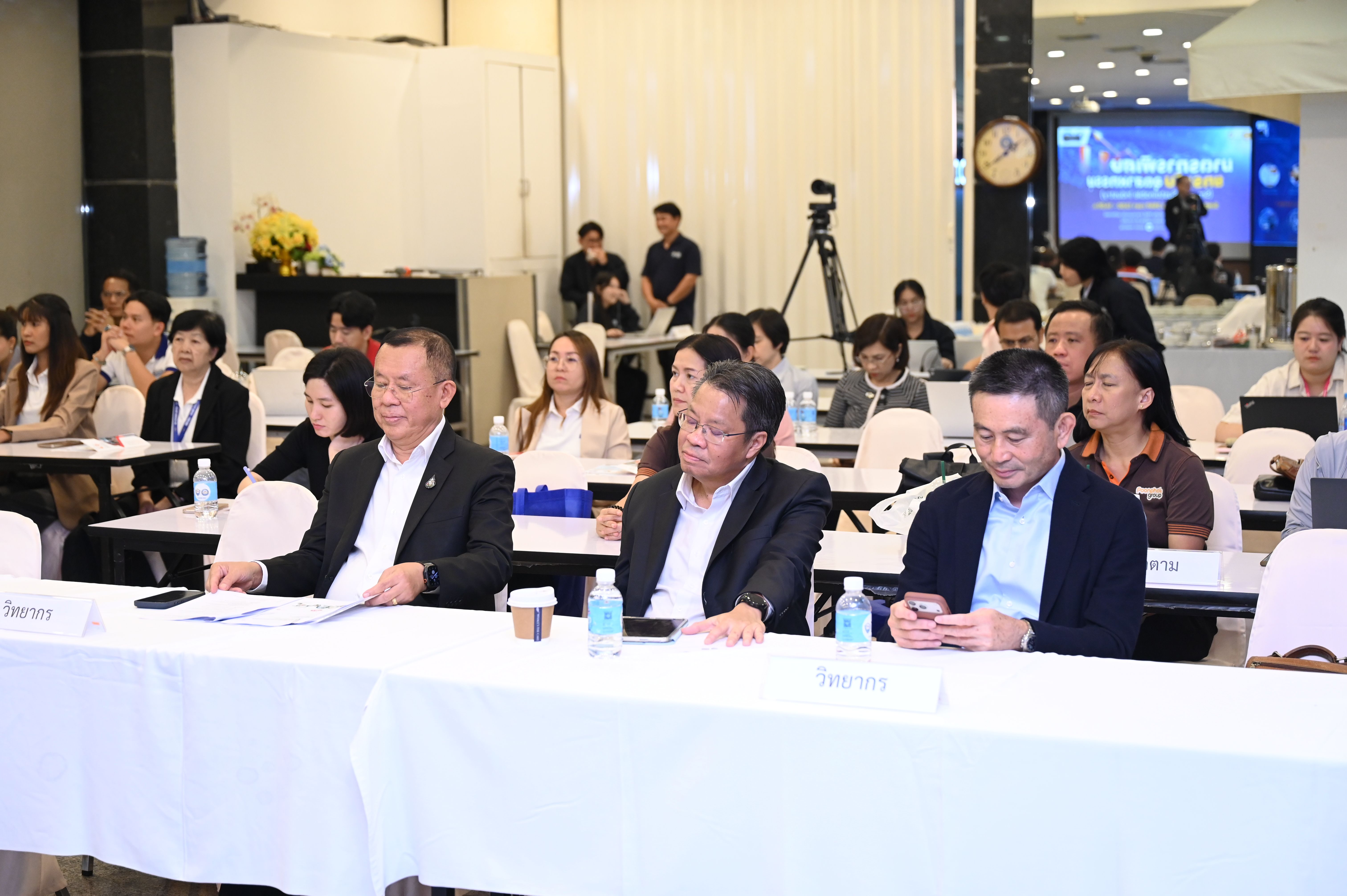 Three businessmen sitting at a table during a conference with other attendees in the background.