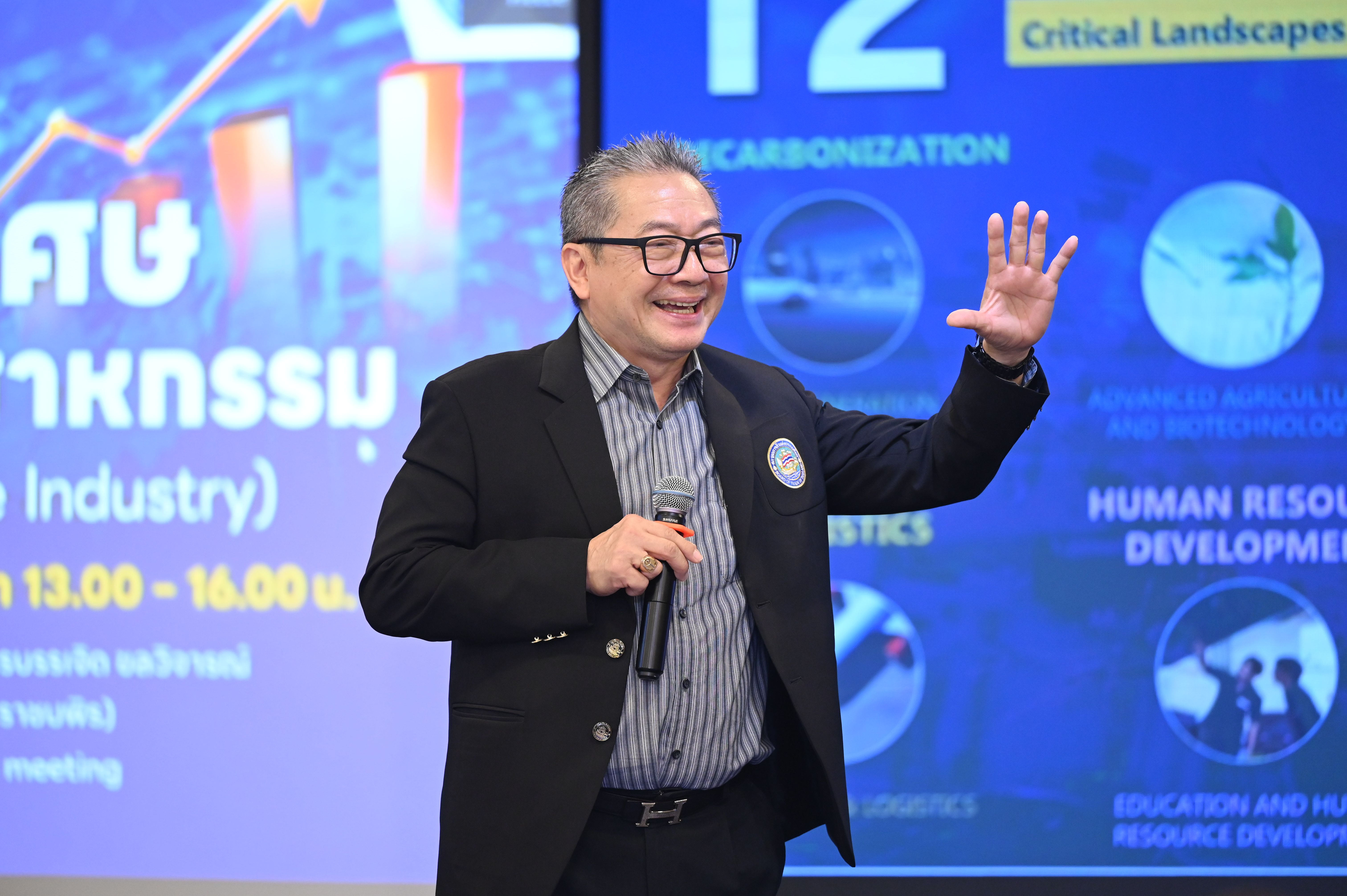 A smiling speaker in glasses presents at a conference, gesturing toward a screen with text.