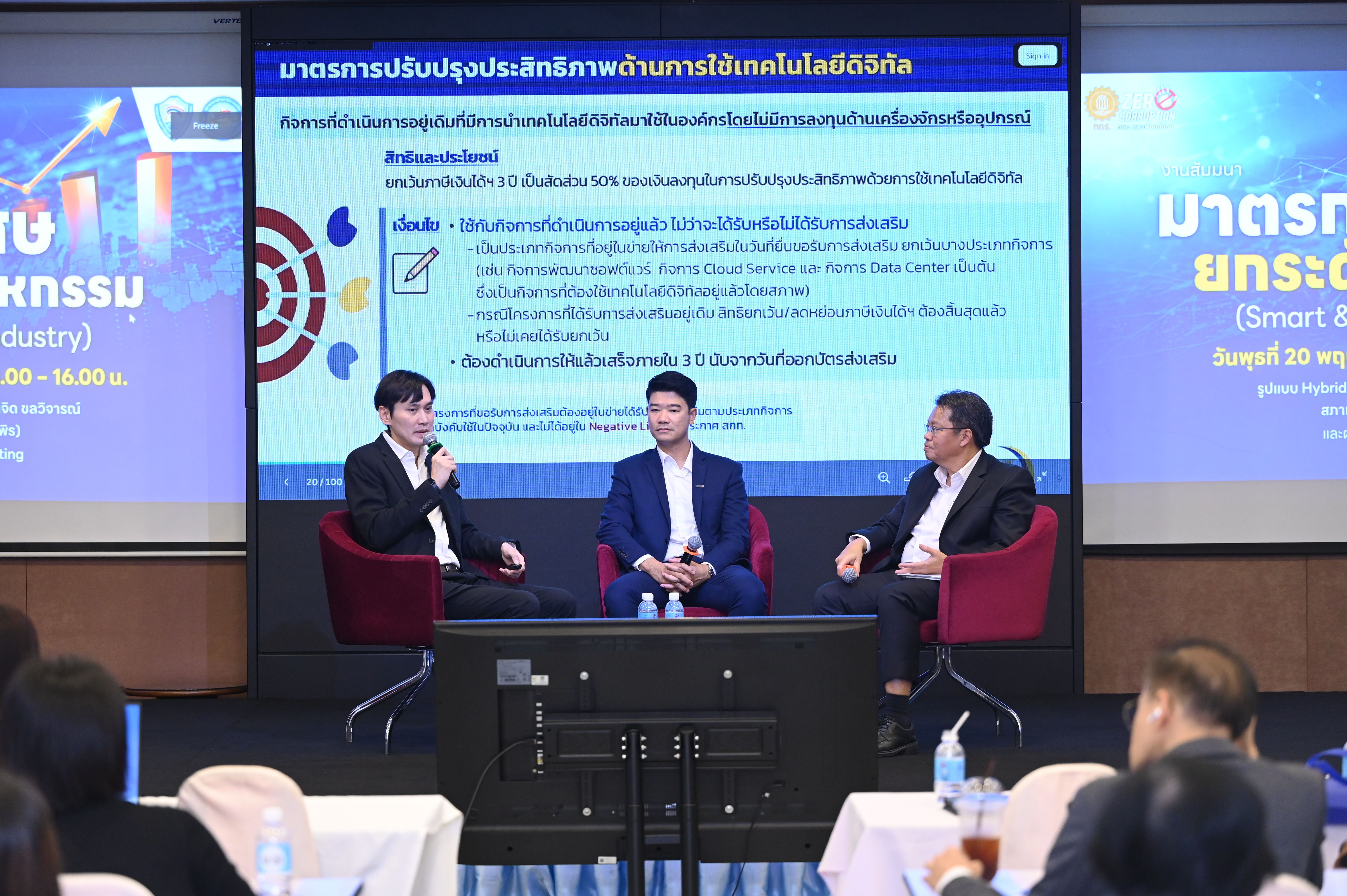 Three men on stage in a panel discussion, a large screen displays Thai text.