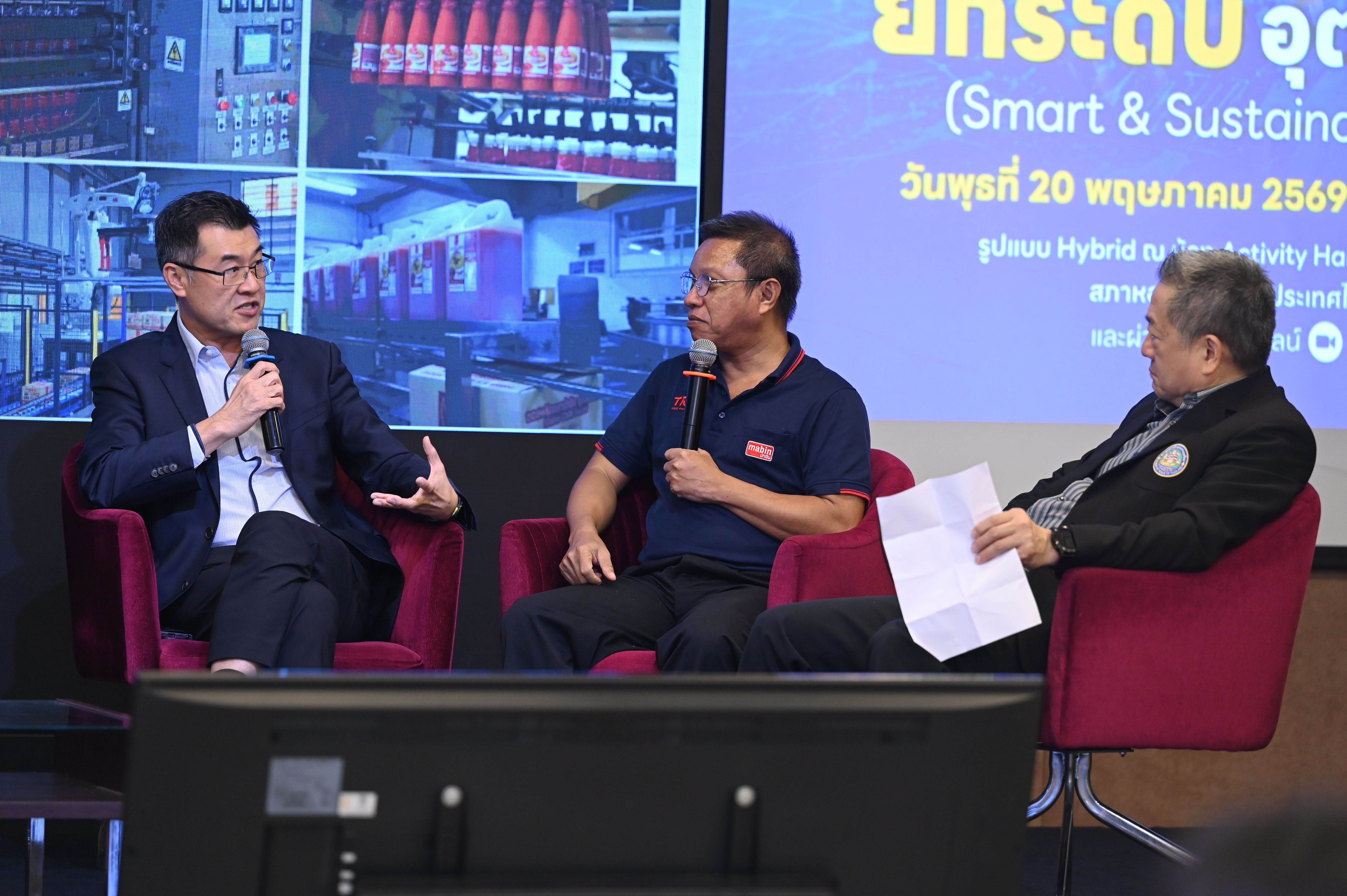 Three men on a panel discussion, two with microphones, seated in front of a digital screen.