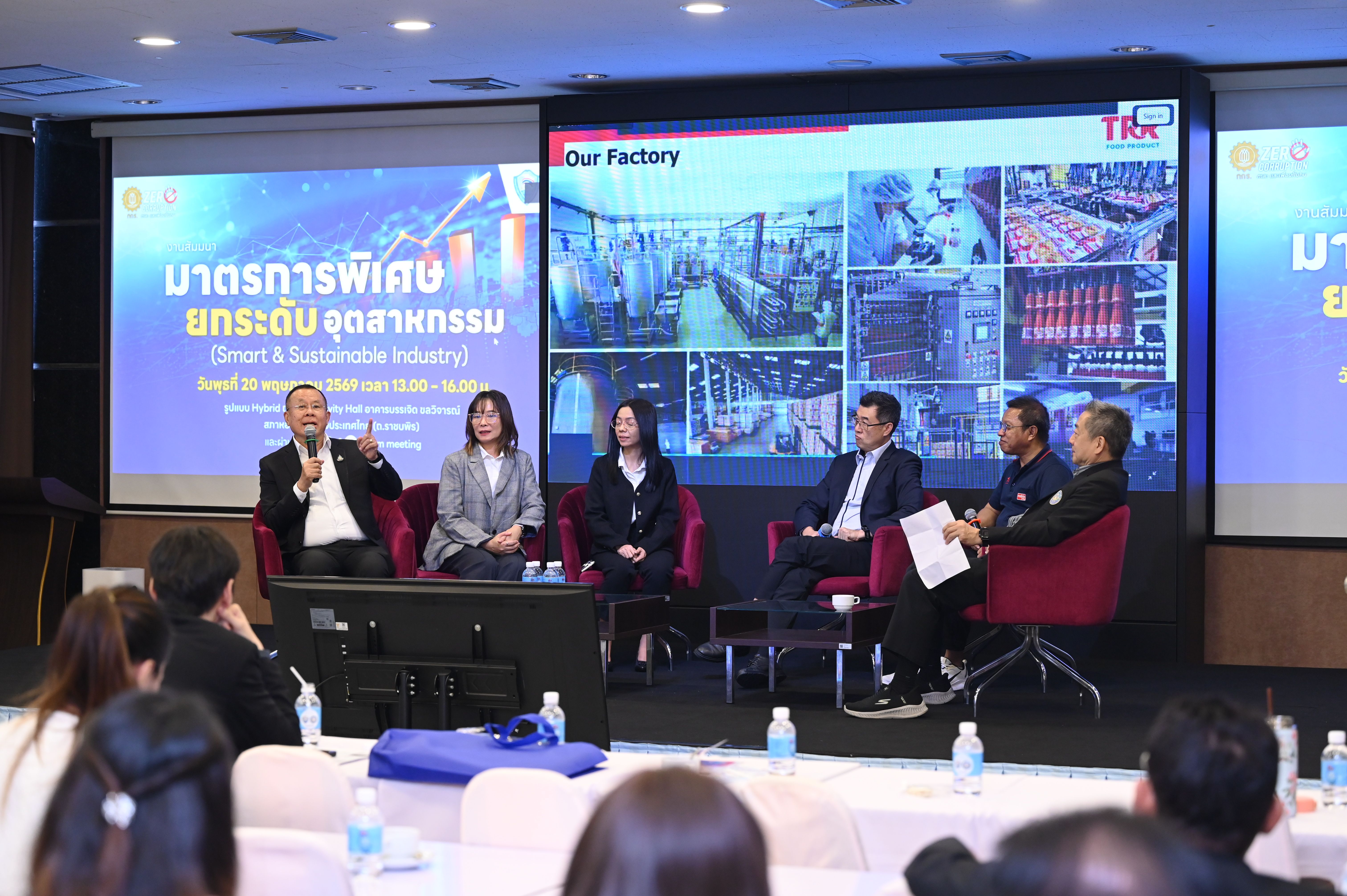 Five panelists on stage discuss industry topics before a large screen displaying factories.
