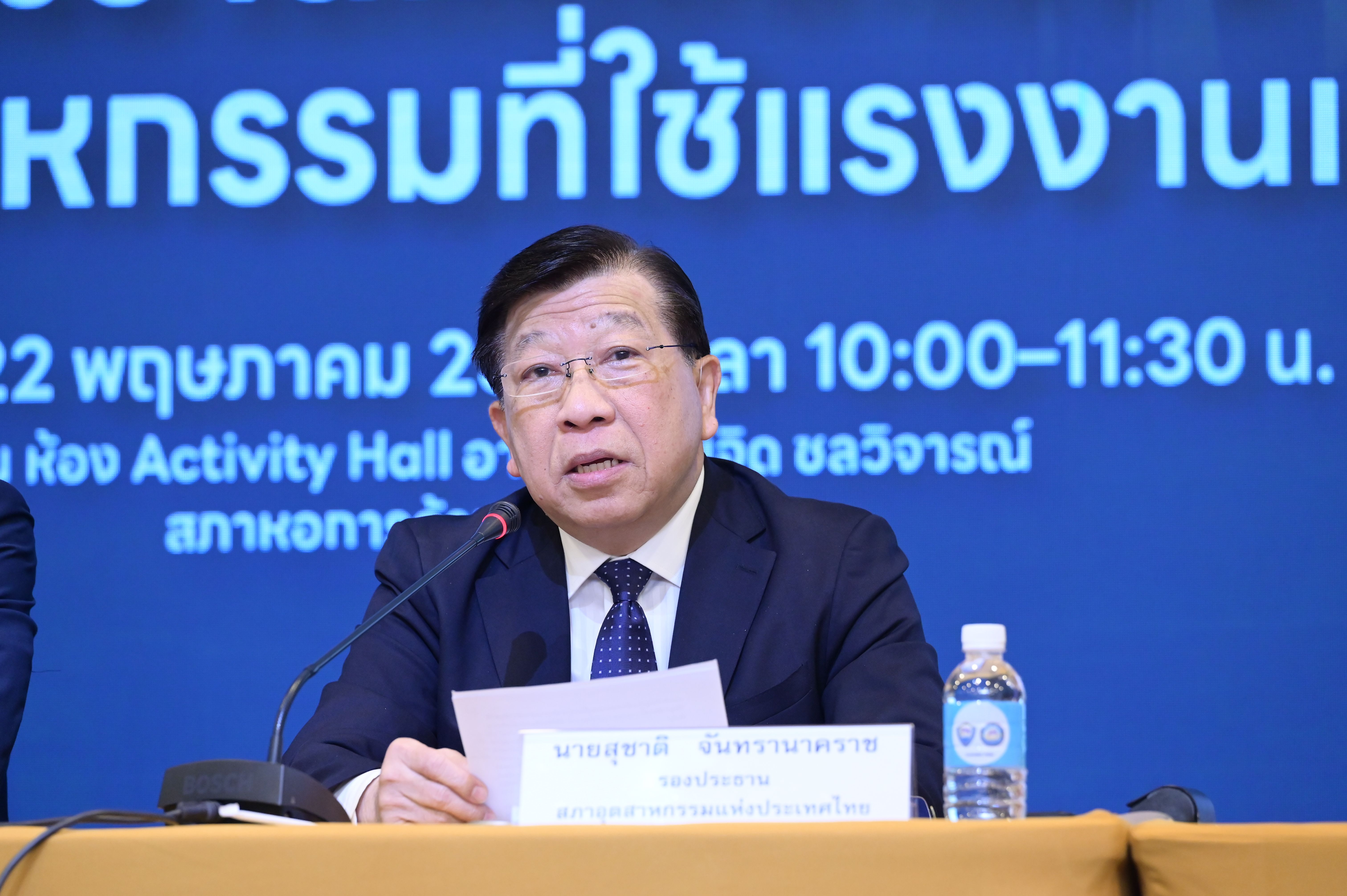 Man speaks at conference podium with Thai text backdrop.