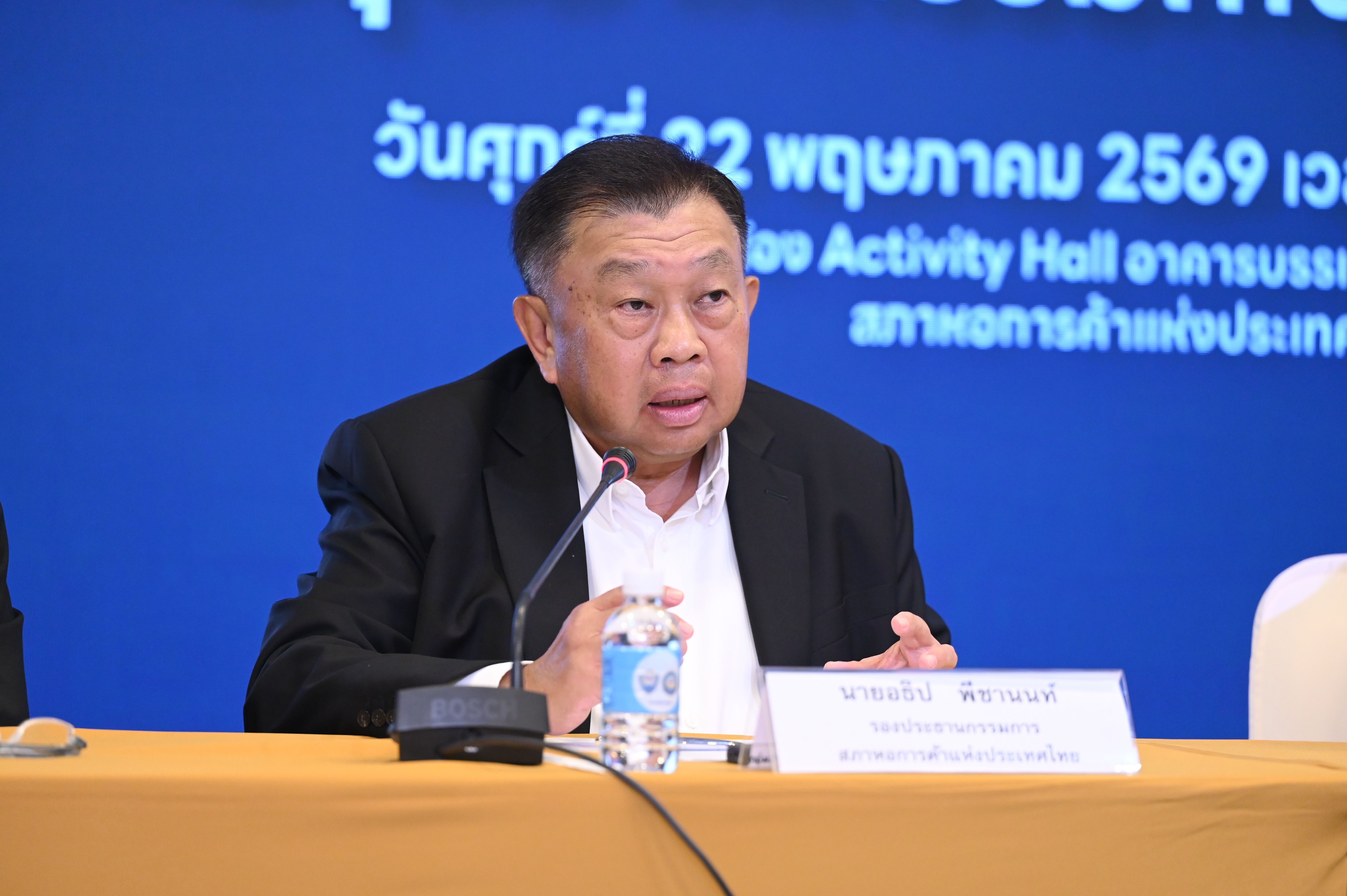 Man in black jacket speaking at a conference table with a microphone and water bottle.