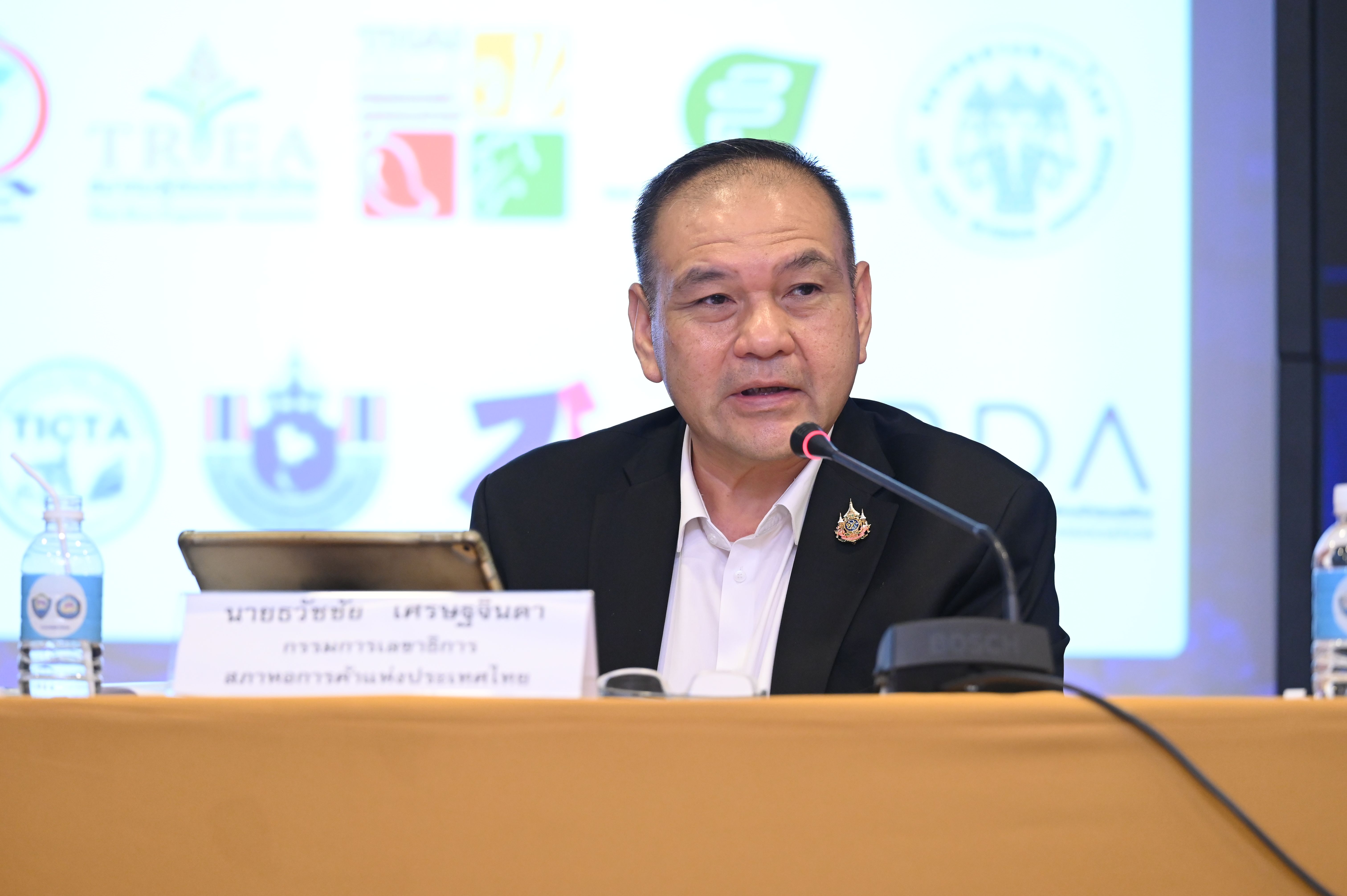 Man in dark suit speaking into a microphone at a formal event with logos behind him.