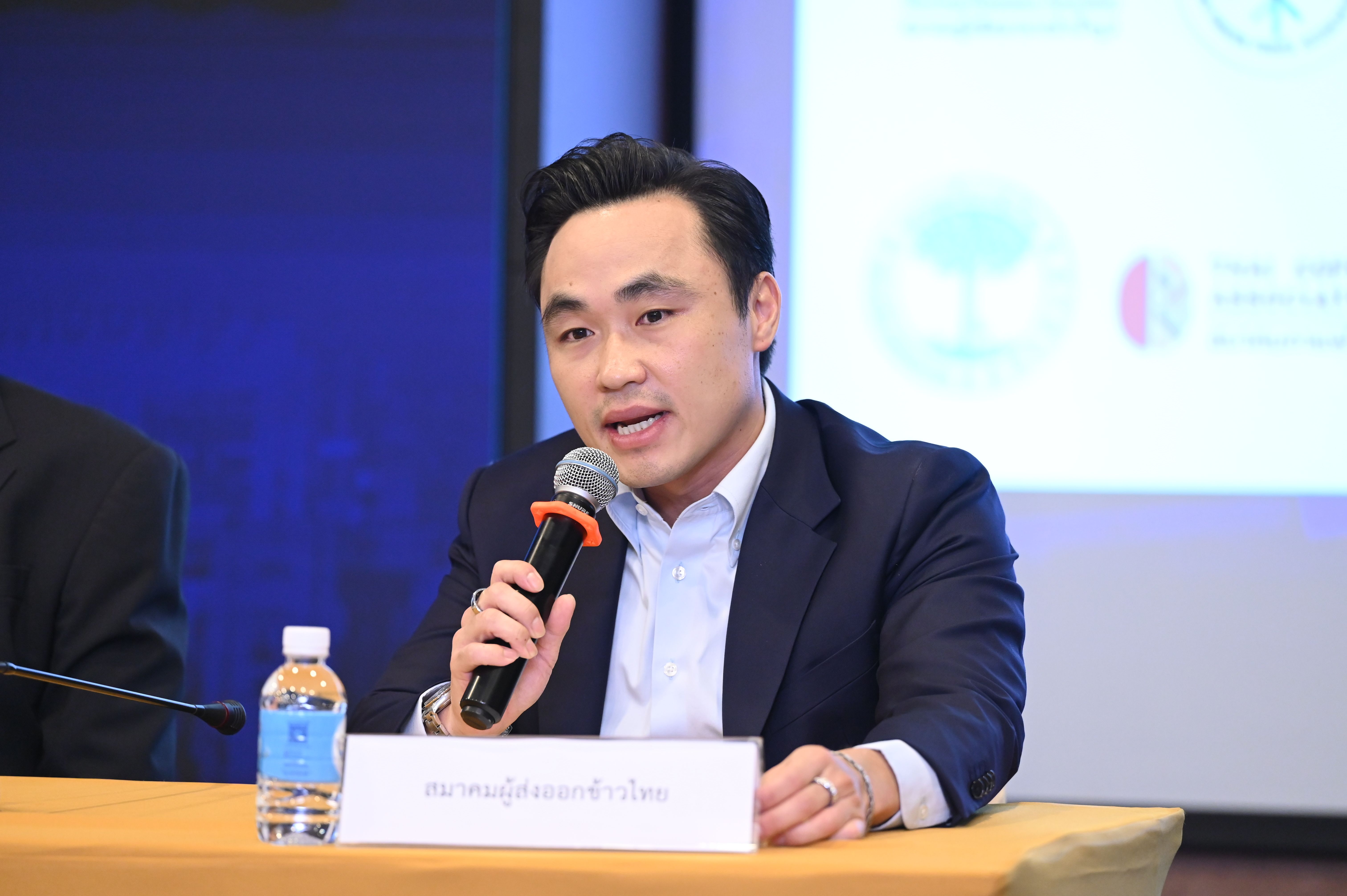 Man in dark suit speaking into a microphone at a conference table.