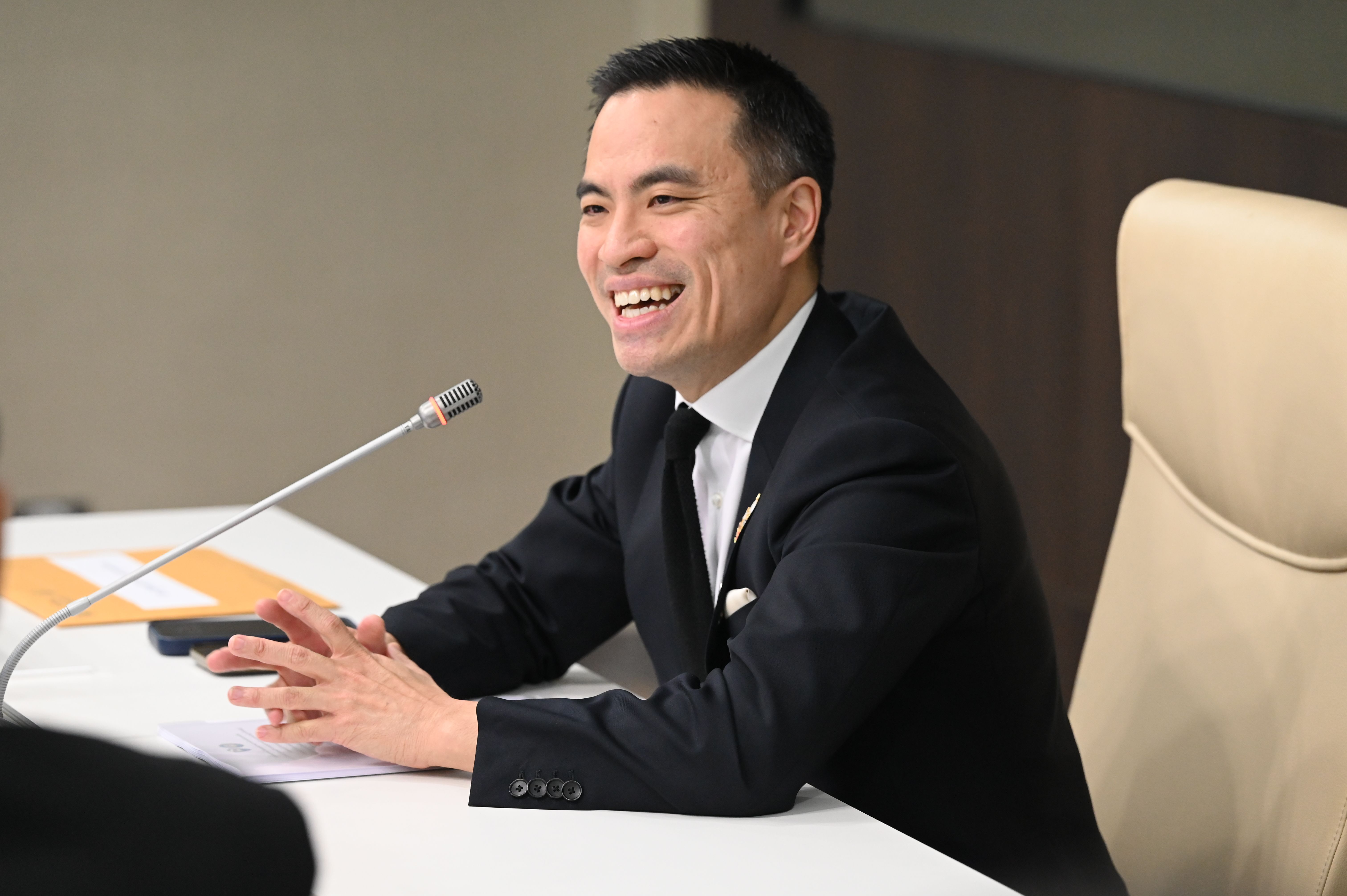 Smiling man in dark suit at a meeting table with a microphone, hands clasped.