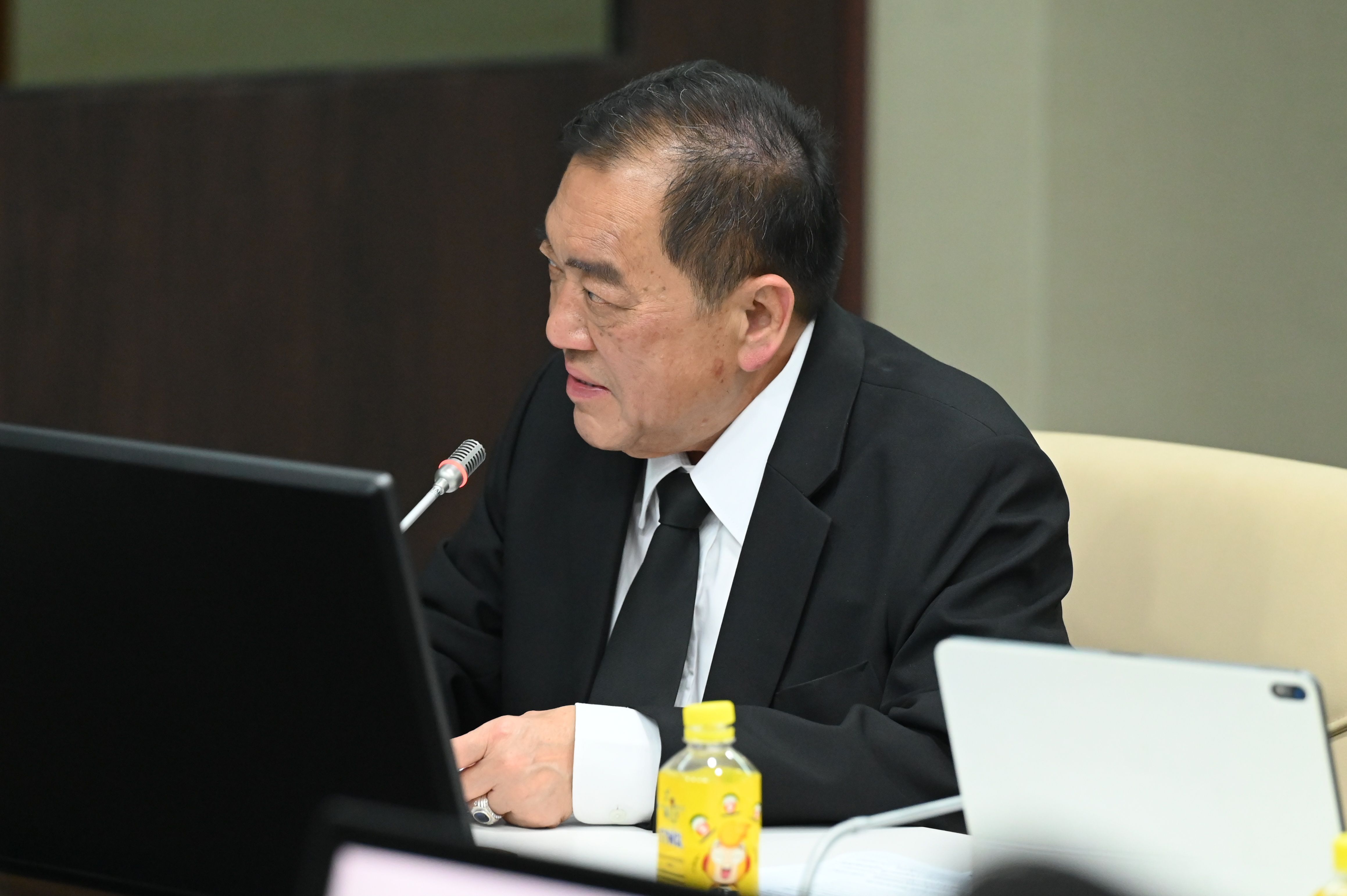 Man in dark suit and tie at a table, focused on a laptop during a meeting.