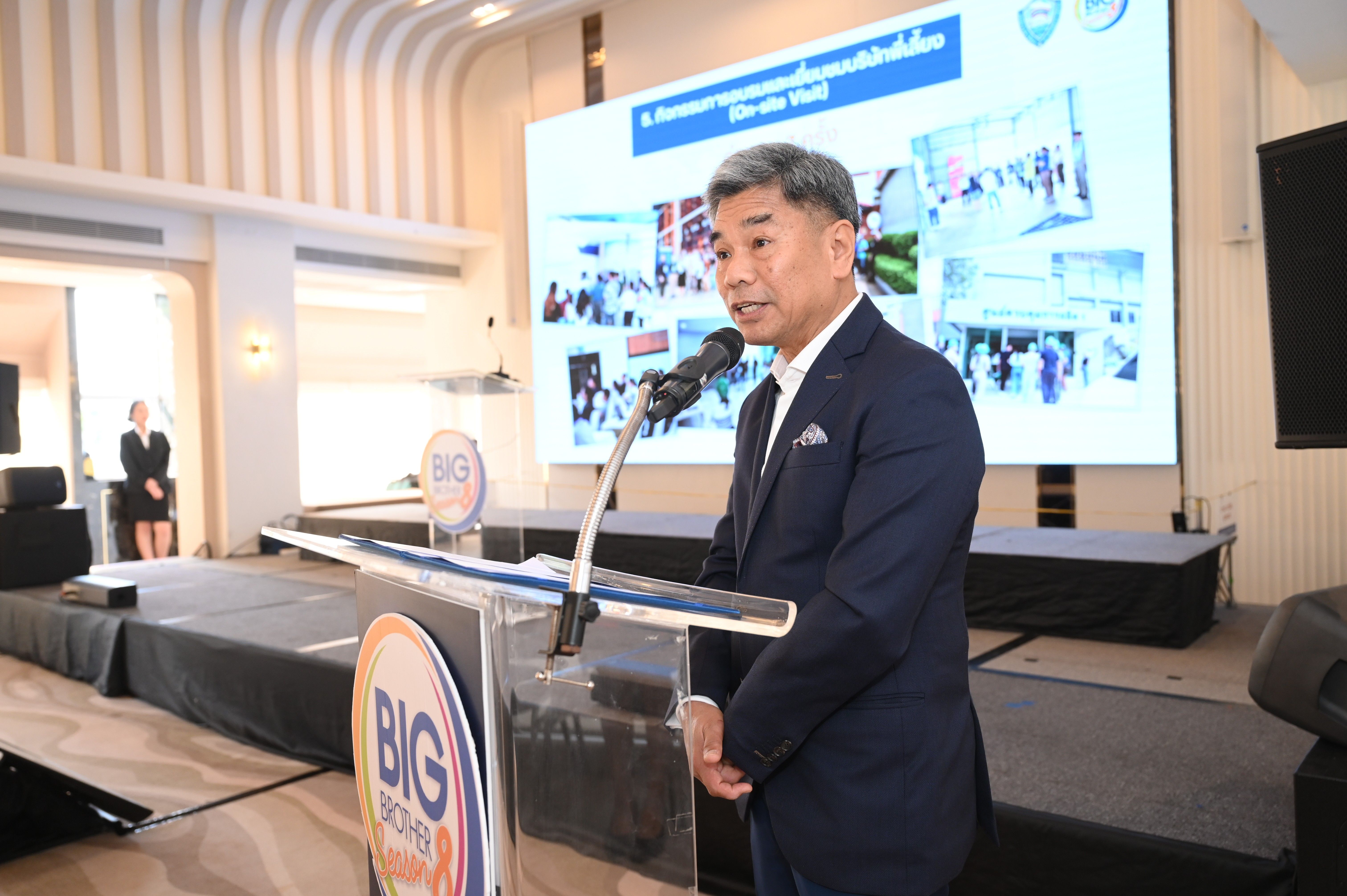 Man in a dark suit speaks at a podium with a large presentation screen behind him.