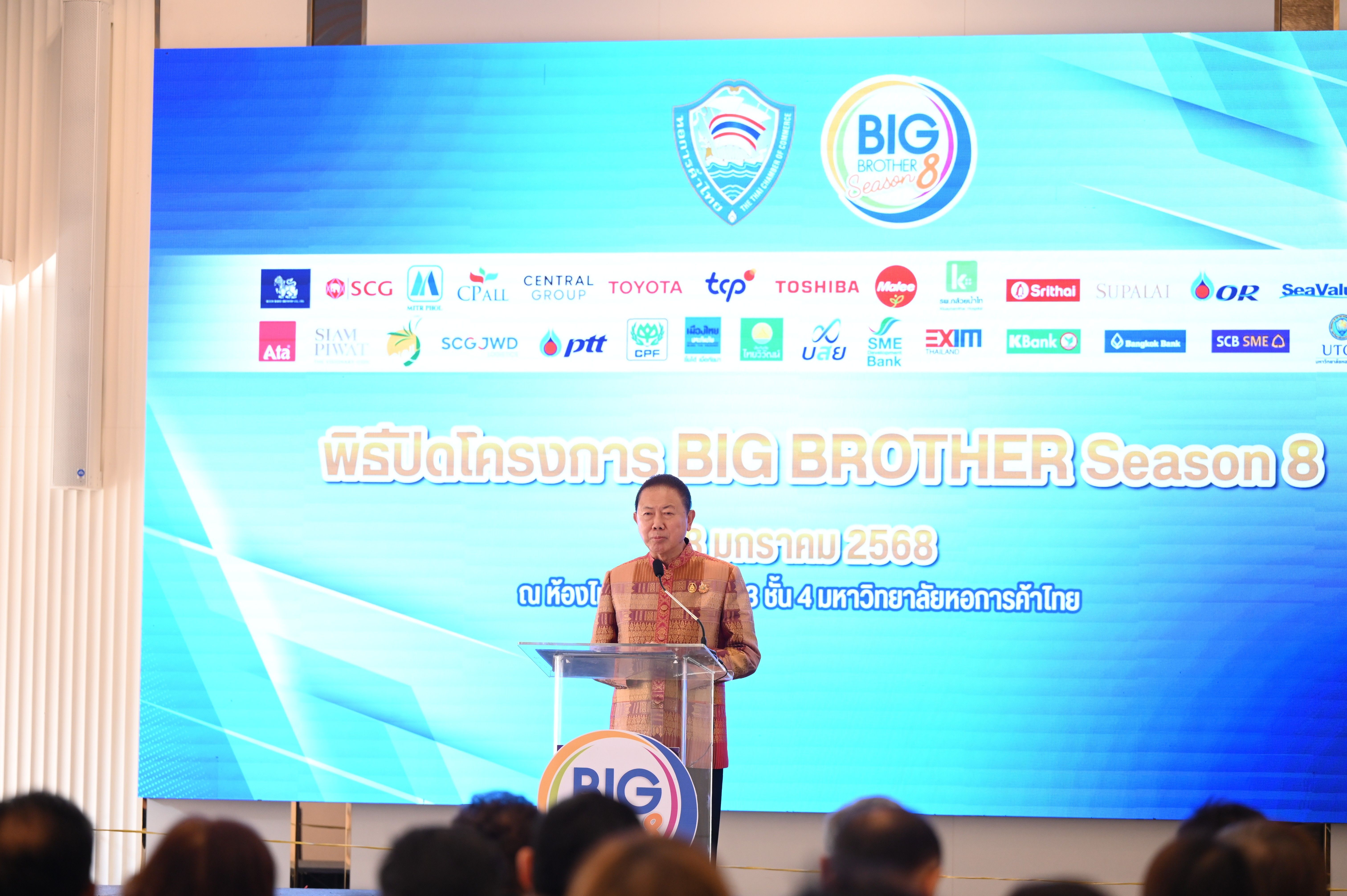 Man speaking at a podium before a large screen displaying "BIG BROTHER Season 8" and logos.