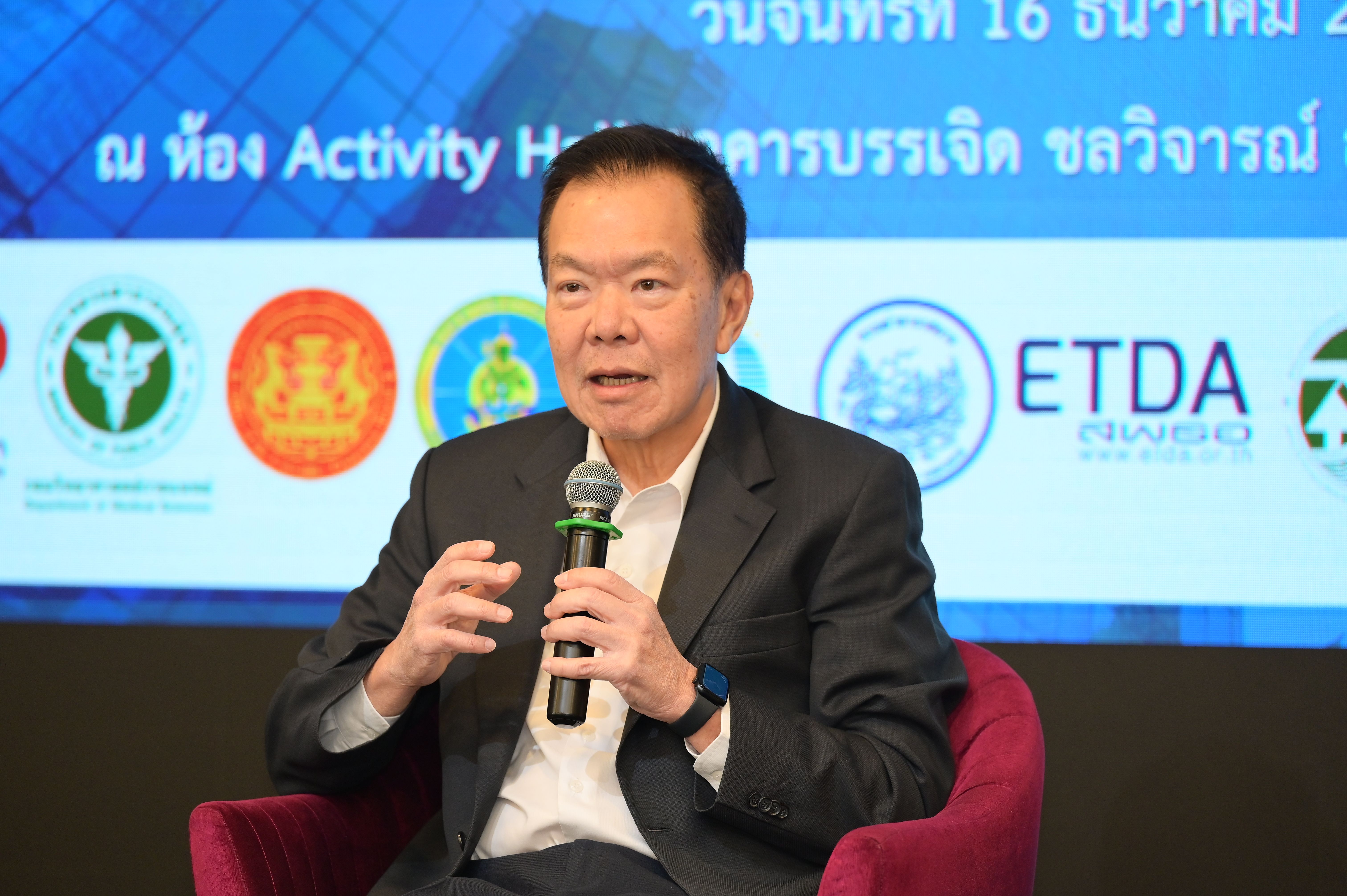 An Asian man in a dark suit speaks into a microphone at a professional event.