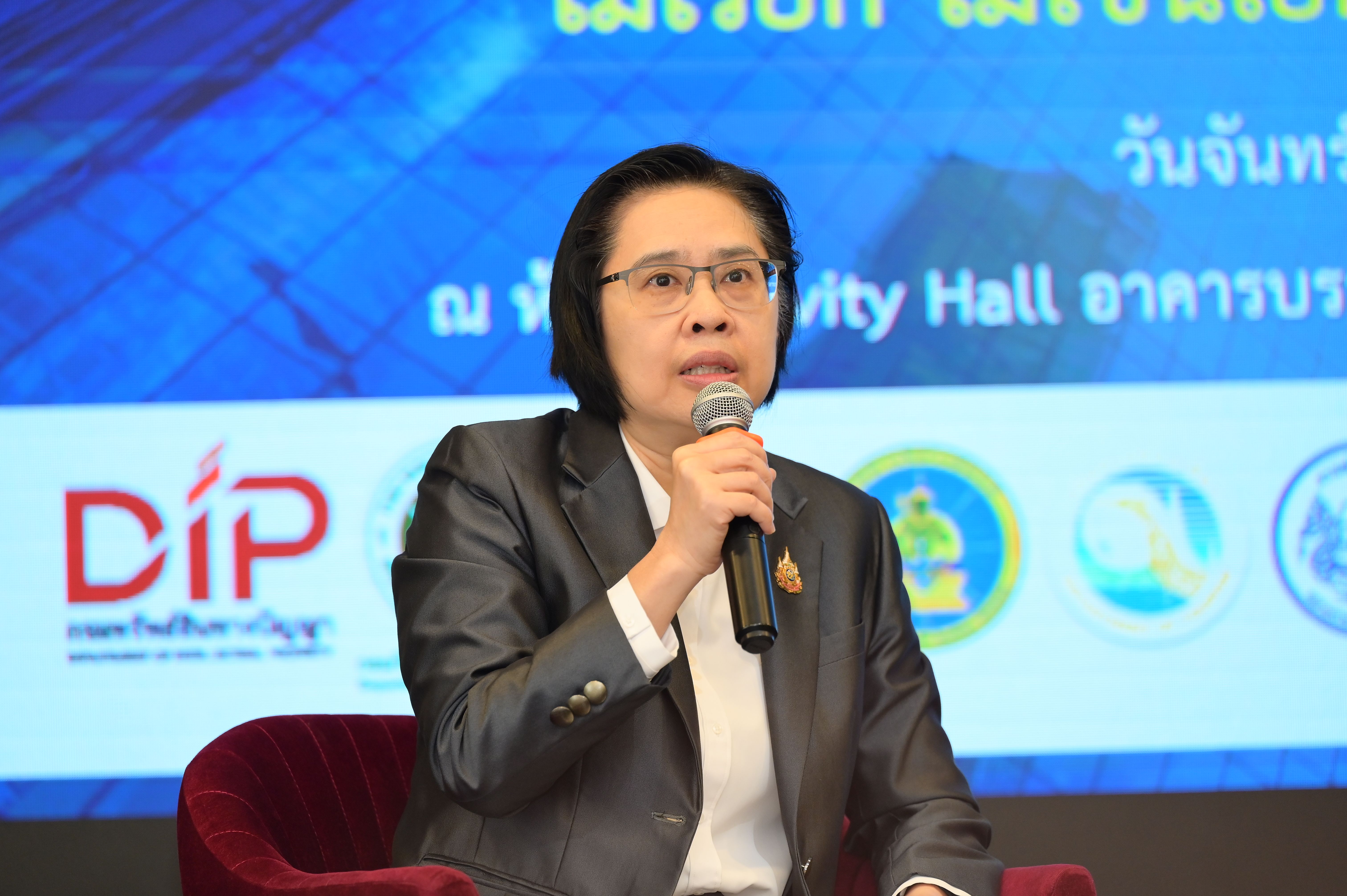Woman in glasses and suit speaking into a microphone at a professional event.