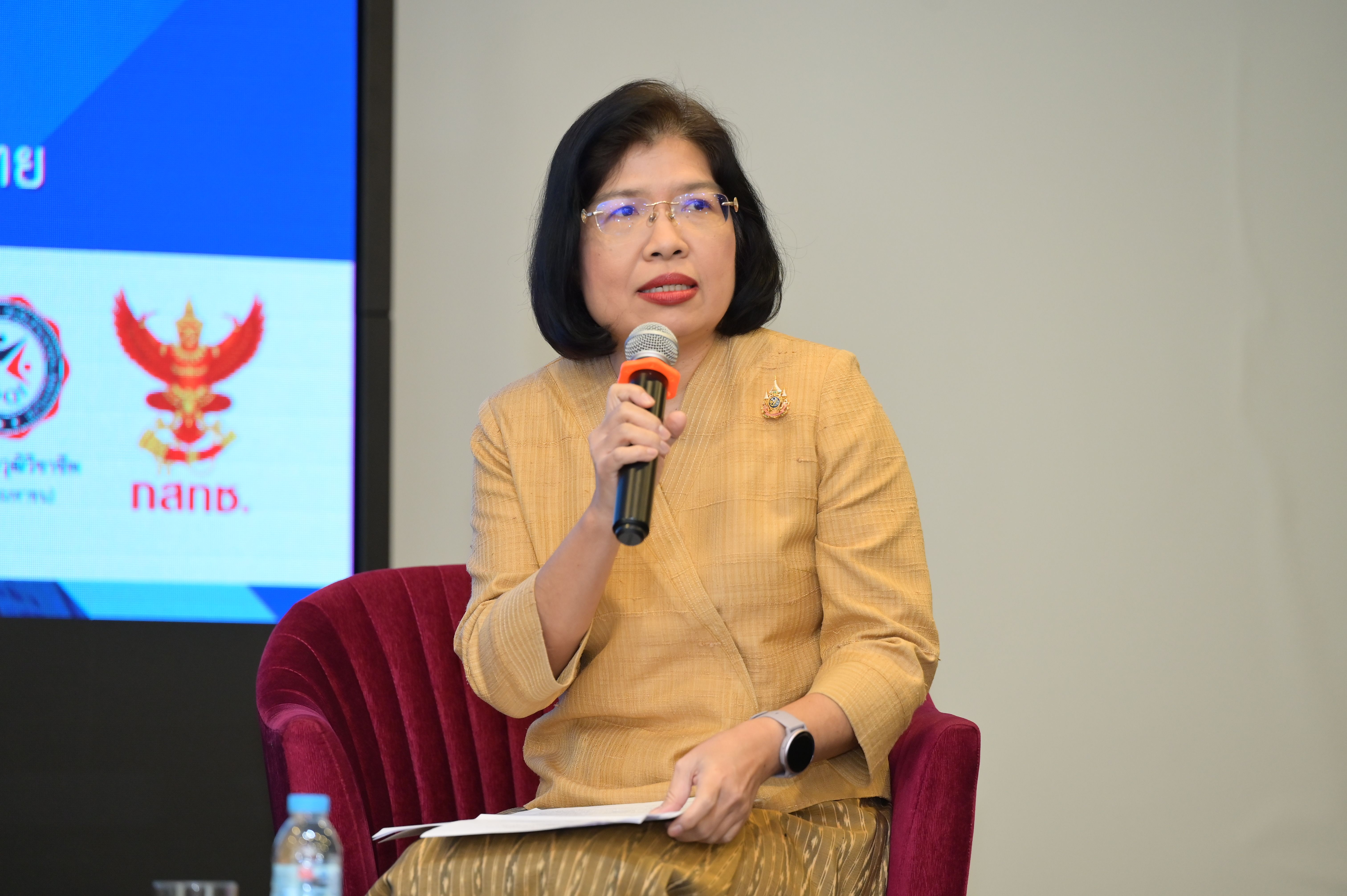 A bespectacled woman in a gold traditional top speaks into a microphone.