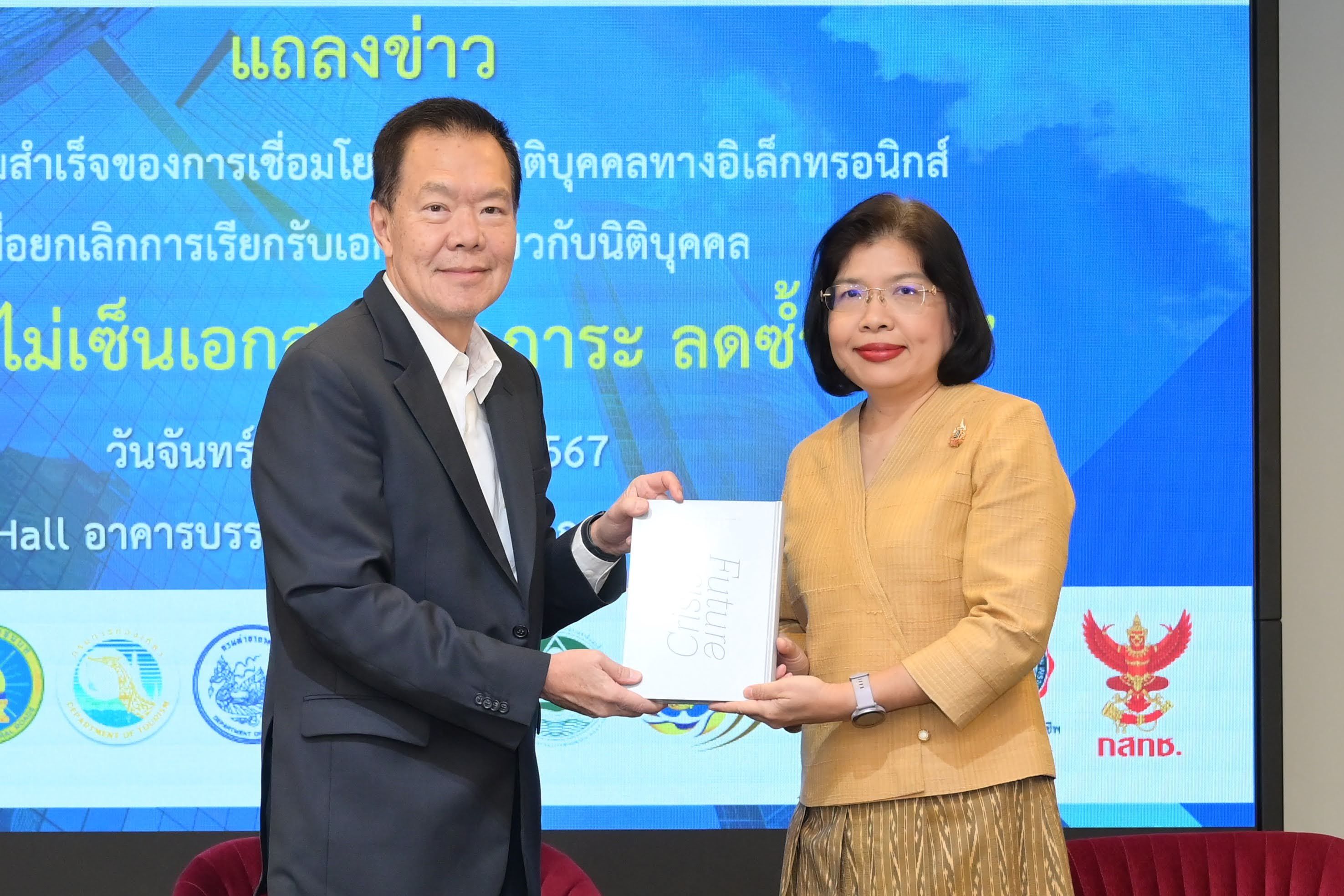 A man and woman exchange a document at a press conference with Thai text backdrop.
