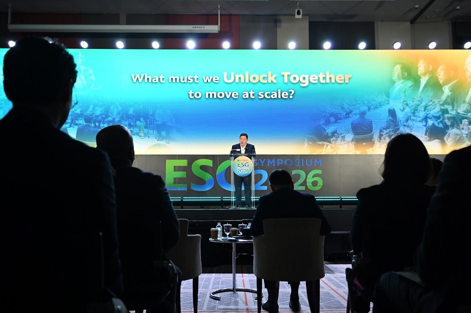 Speaker on stage addressing an audience, screen displays 'Unlock Together' and 'ESG 26'.