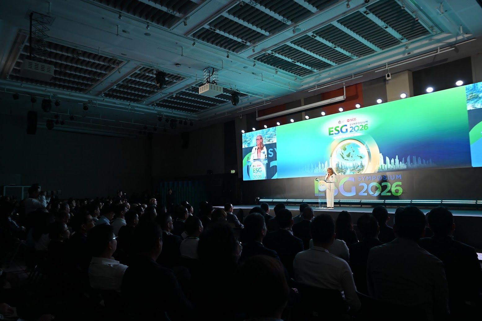 Speaker presents on stage, large screen displays "G2C26" to a seated audience.