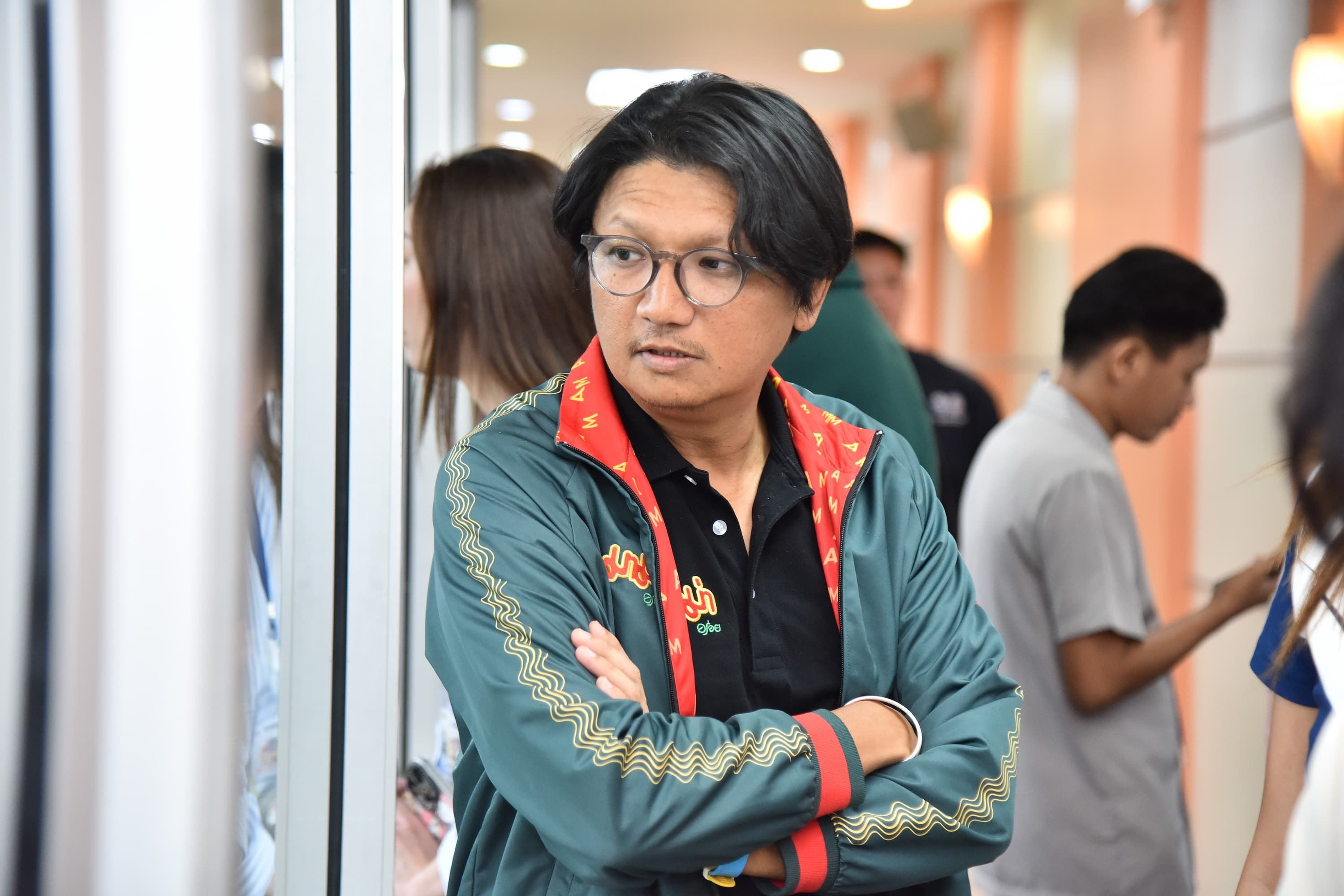 Man with glasses and green jacket, arms crossed, looking thoughtfully to the side.
