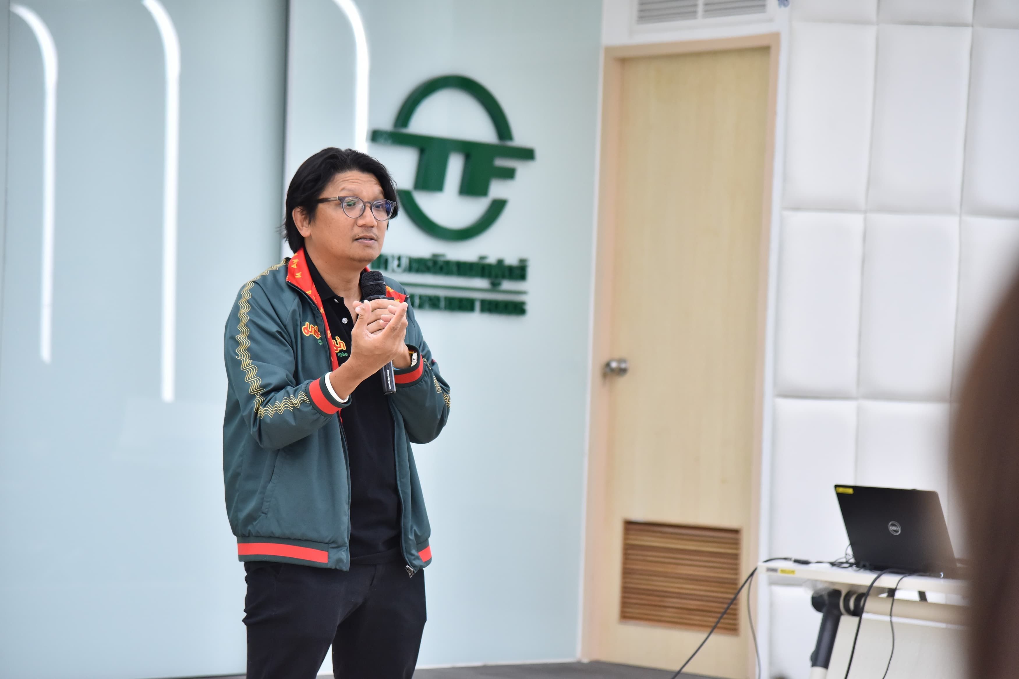 Man with glasses in a green jacket speaking, hands clasped, in front of a logo.