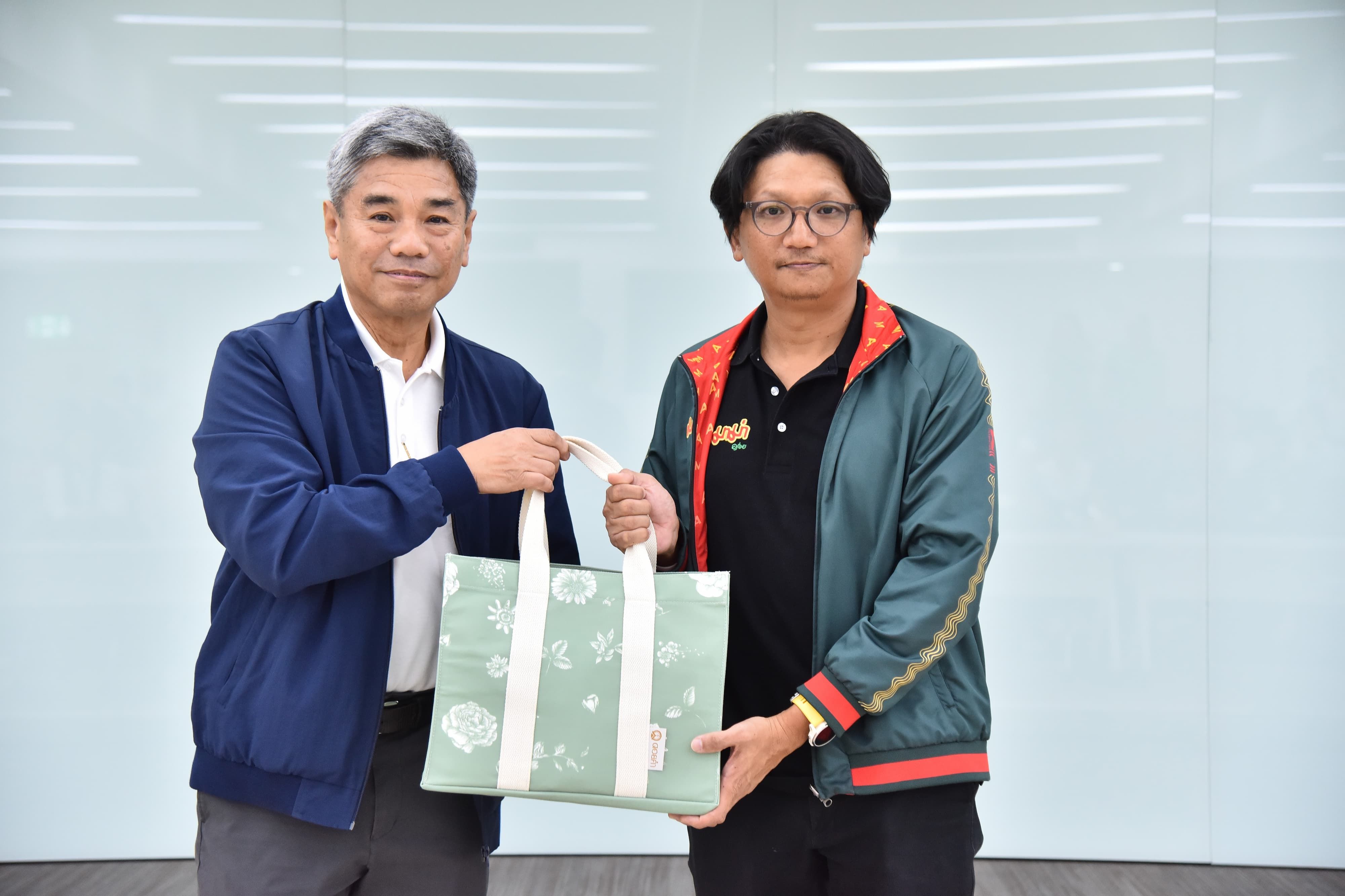 Two men exchanging a light green tote bag in an indoor setting.
