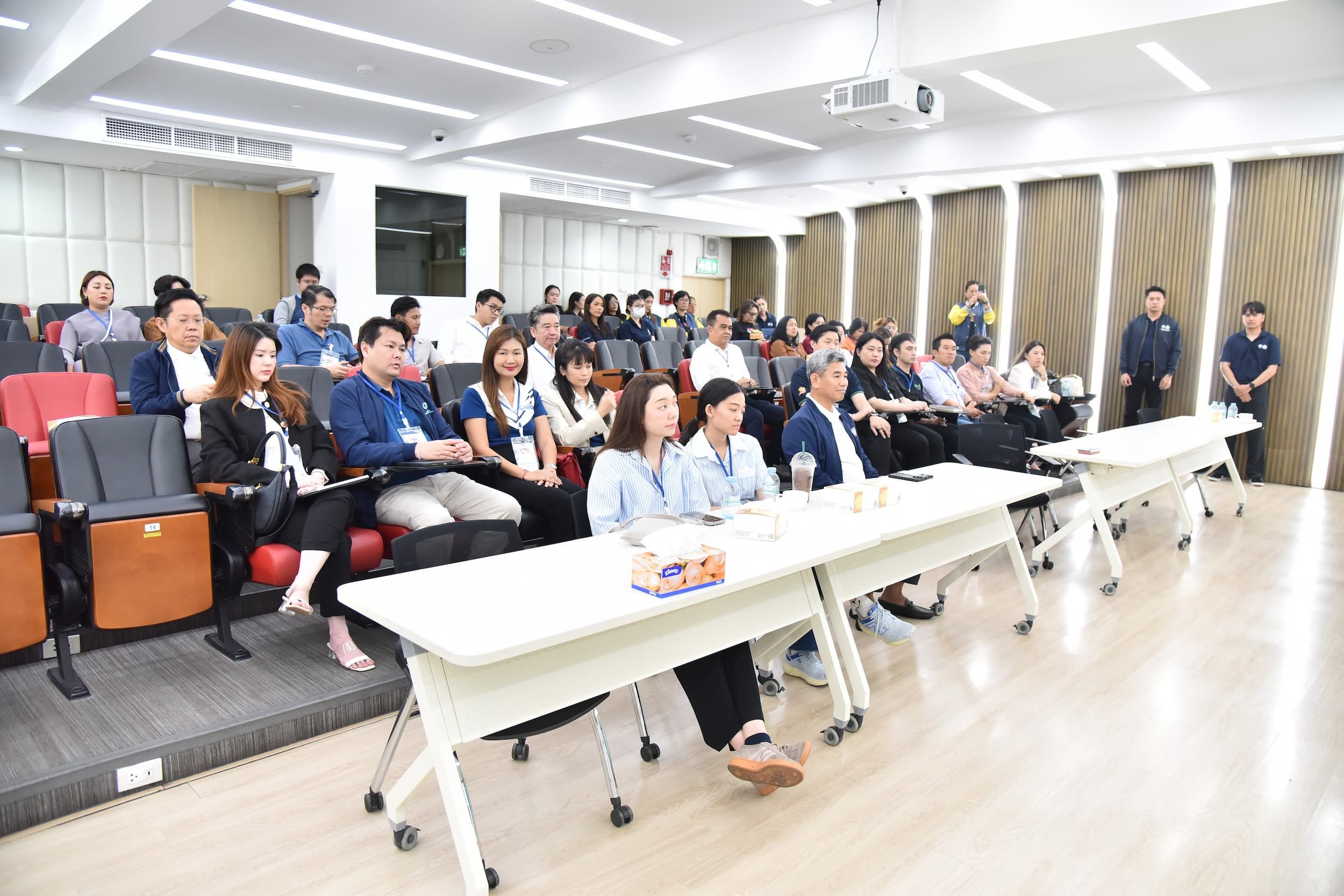 Audience listening attentively in a modern lecture hall with long tables.