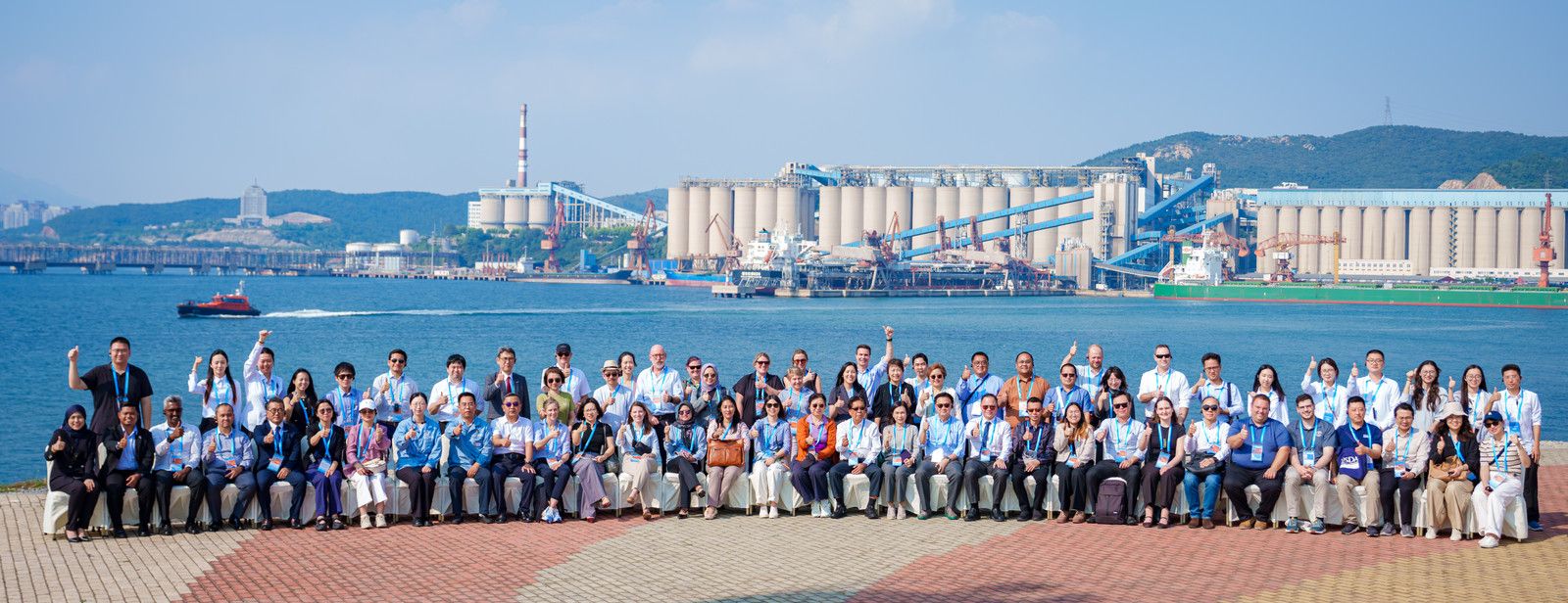 Large group of people posing outdoors by an industrial waterfront with silos.