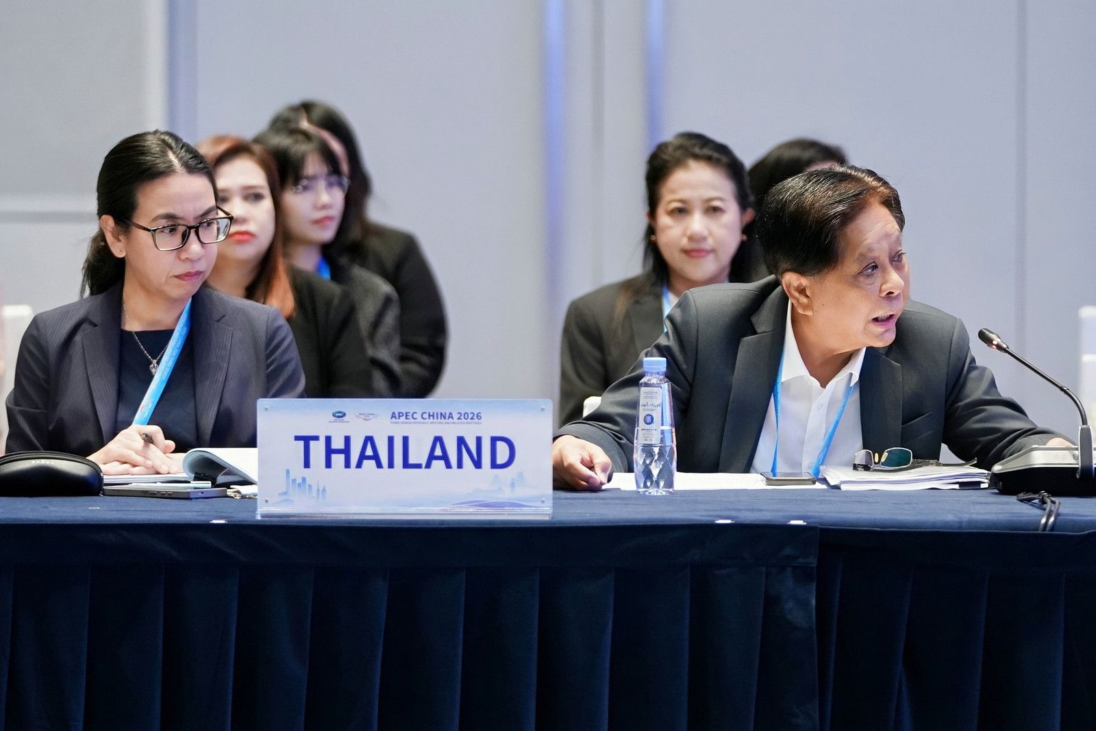 Man speaks at a conference table with a 'THAILAND' nameplate, accompanied by a woman.