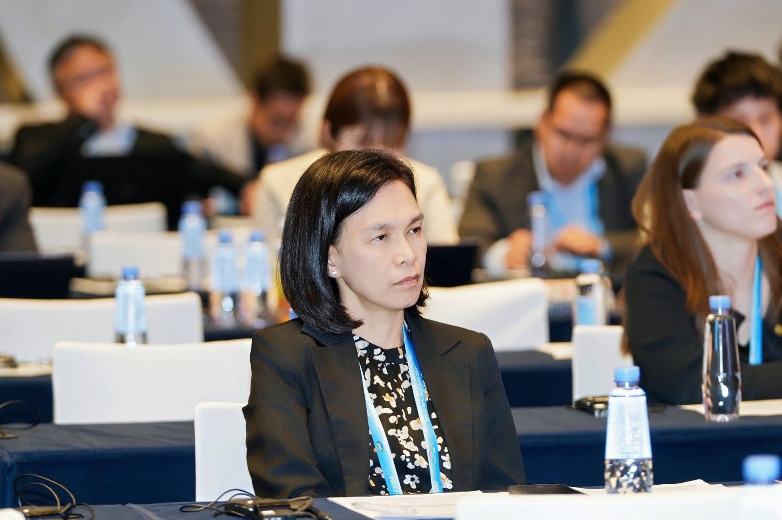 A professional woman with dark hair sits attentively at a conference table.