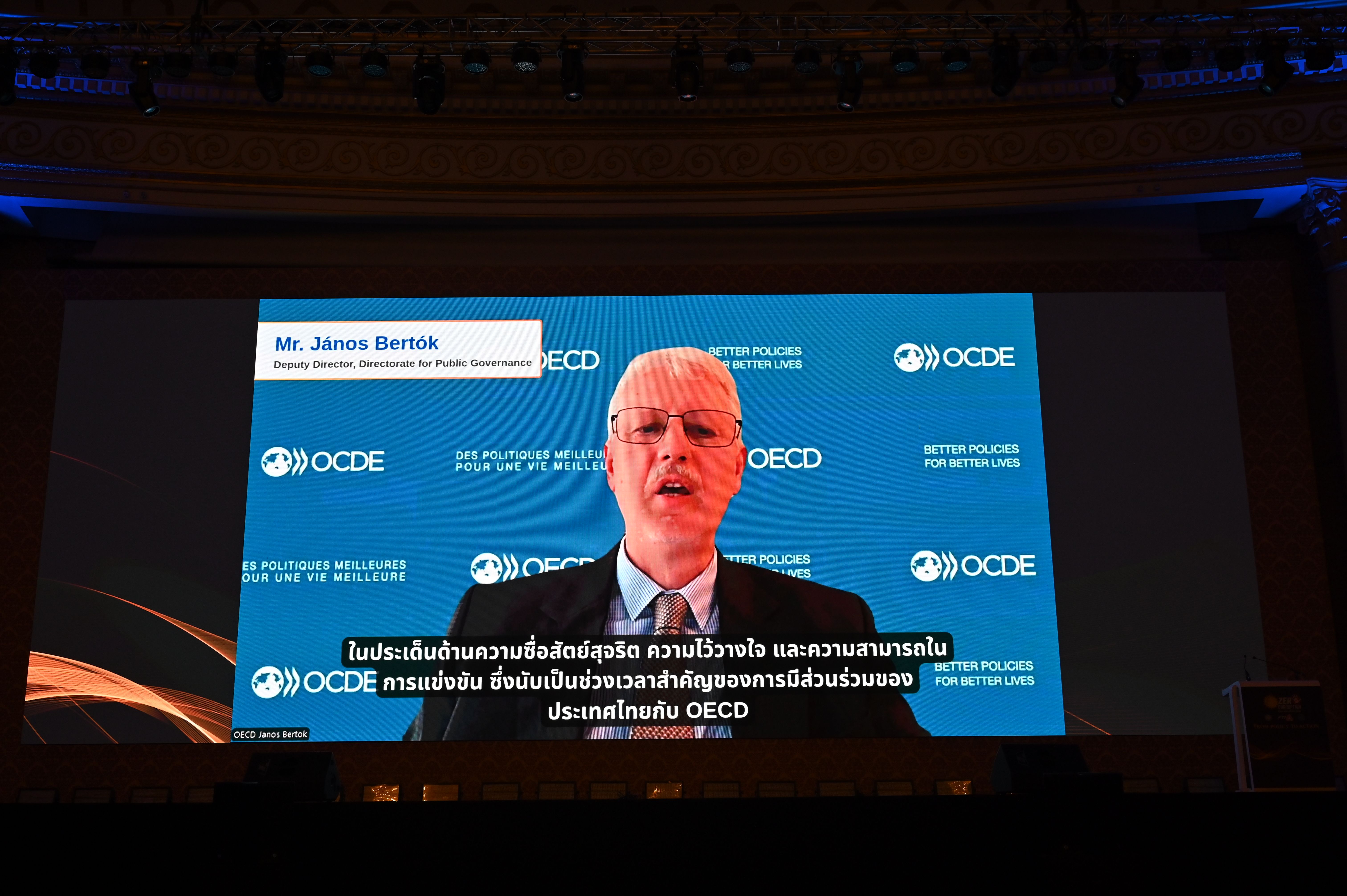 Man speaks on a large screen with OECD logo and foreign subtitles during a presentation.