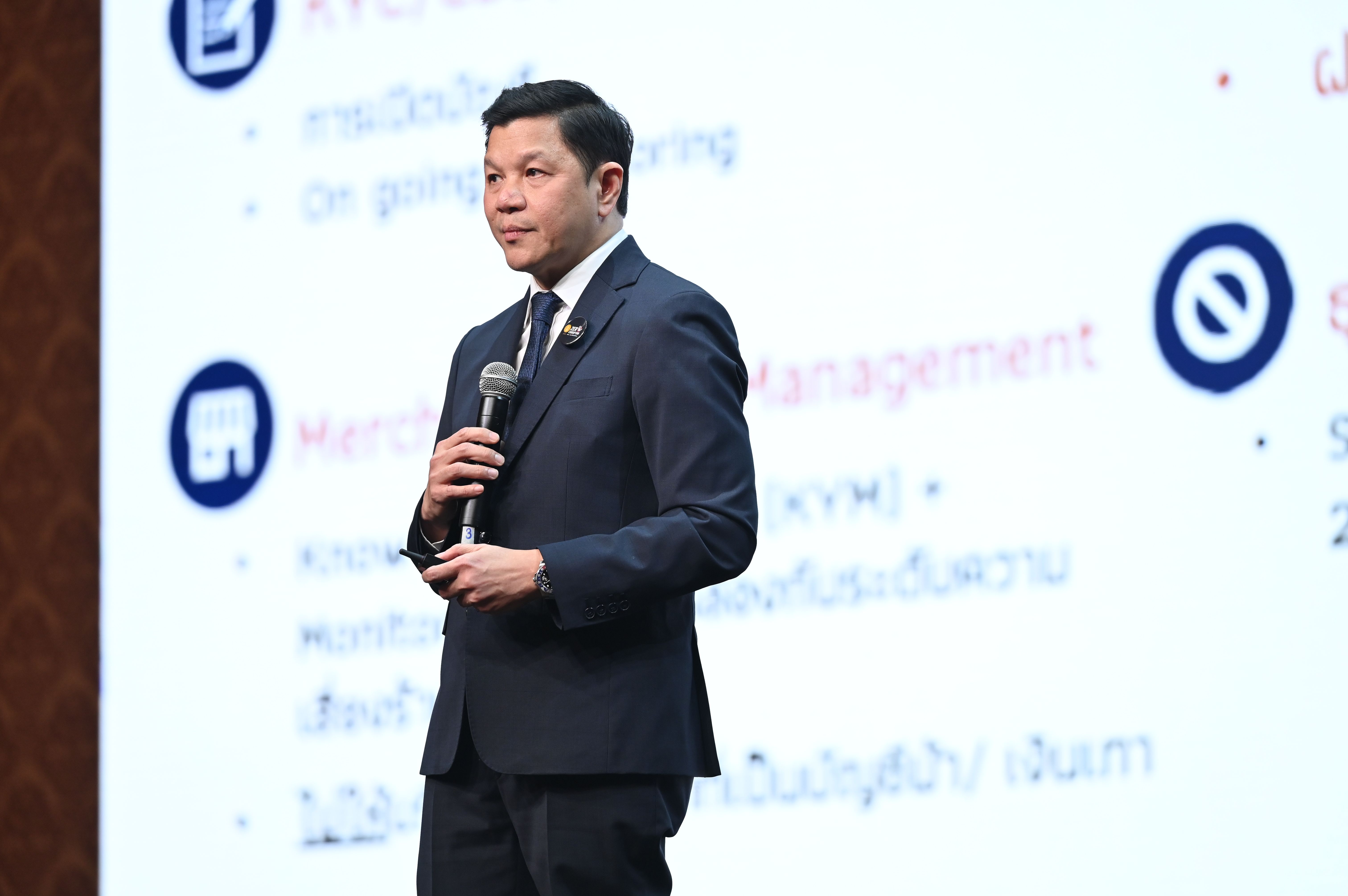 A man in a dark suit speaks into a microphone during a presentation.