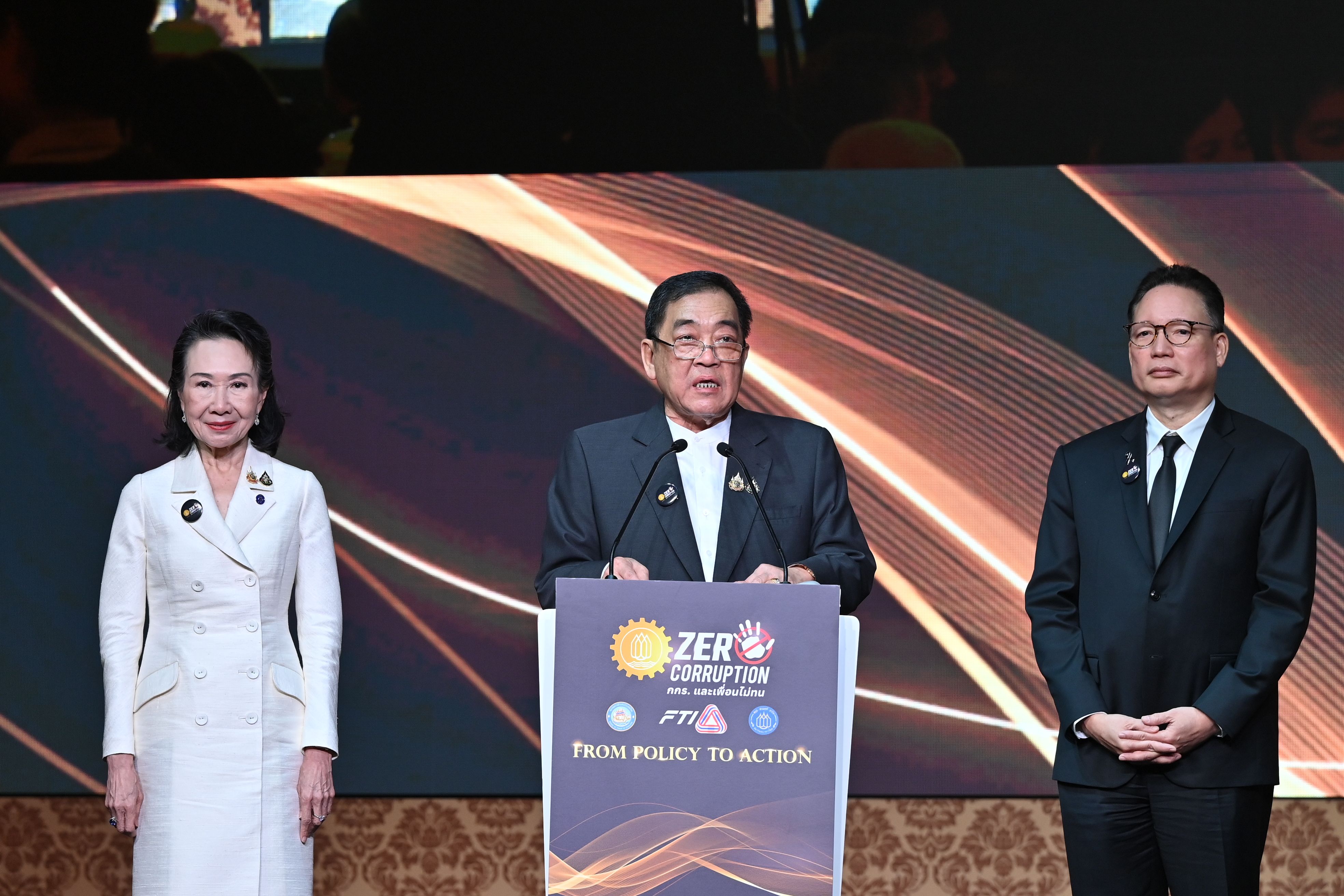 Three people on stage, a man speaking at a podium with "ZERO CORRUPTION" text.