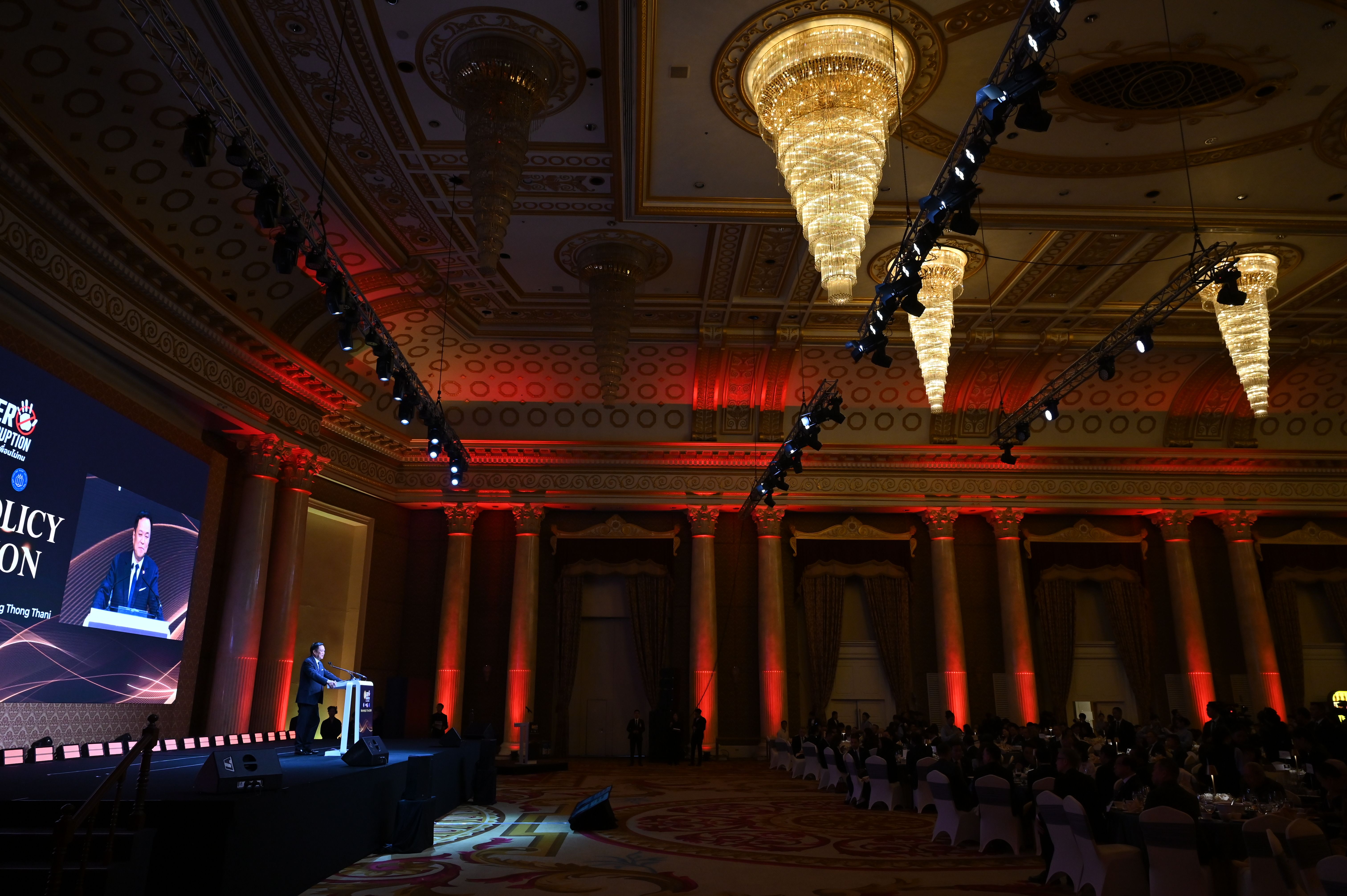 Speaker presents on stage in an opulent conference hall with chandeliers and red lighting.