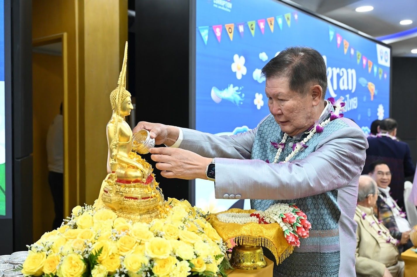 An older man in a suit pours water over a golden Buddha statue.