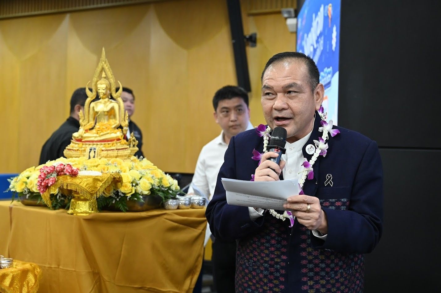 A man speaks into a microphone next to a golden Buddha statue.