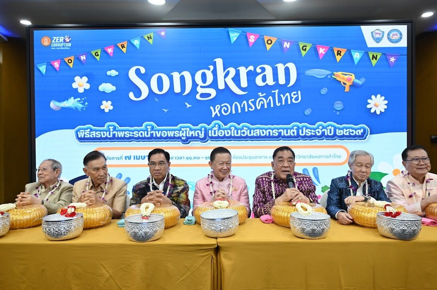 Seven men sit at a table in front of a 'Songkran' event screen.