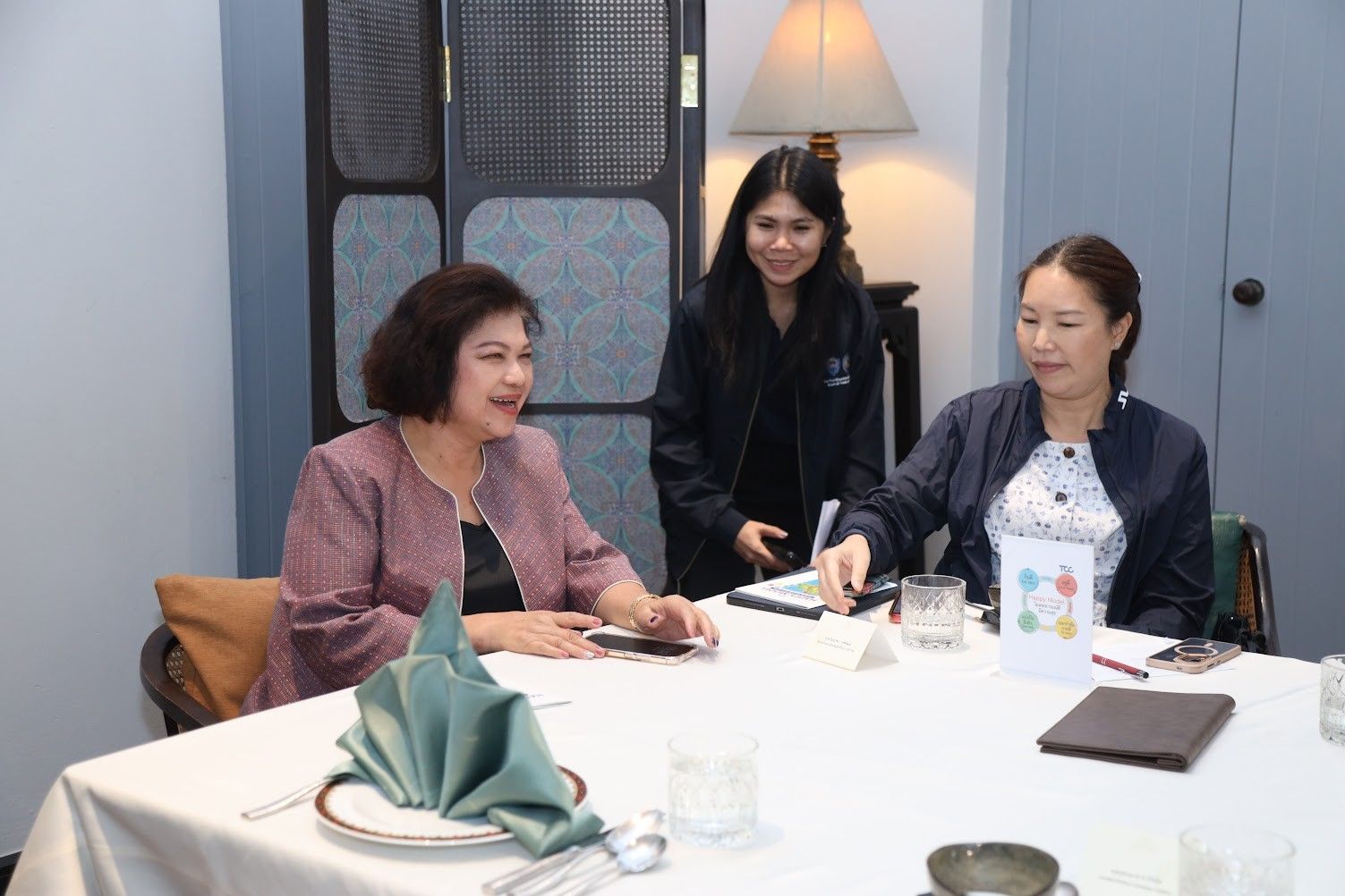 Three women, two seated and one standing, engage in conversation at a restaurant table.