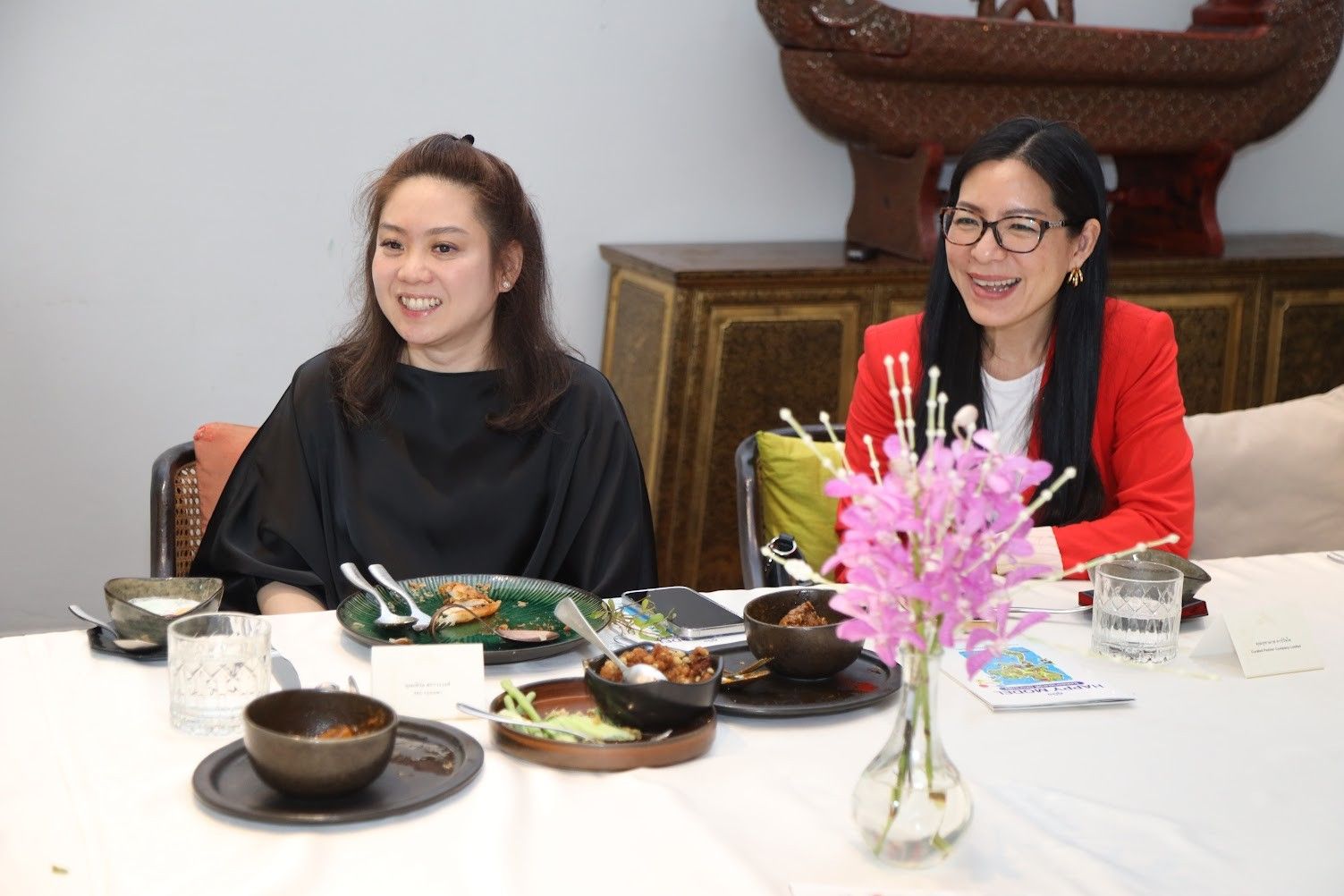 Two smiling women seated at a table with food and a vase of pink flowers.