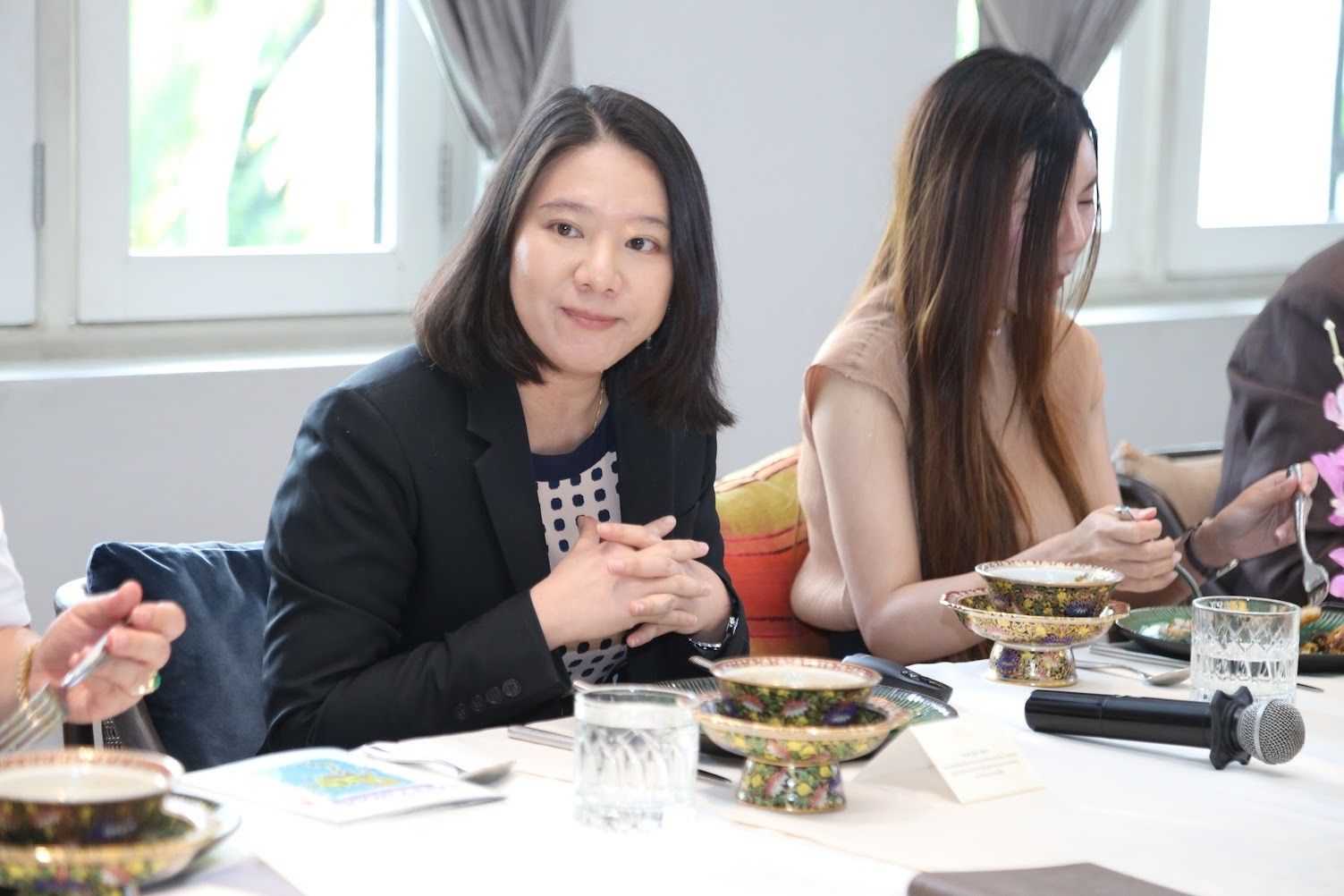 A professional woman with dark hair sits at a table, hands clasped, smiling slightly.