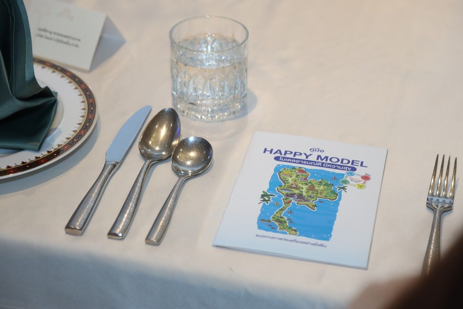 Formal table setting with silverware, water glass, and a brochure titled 'HAPPY MODEL'.