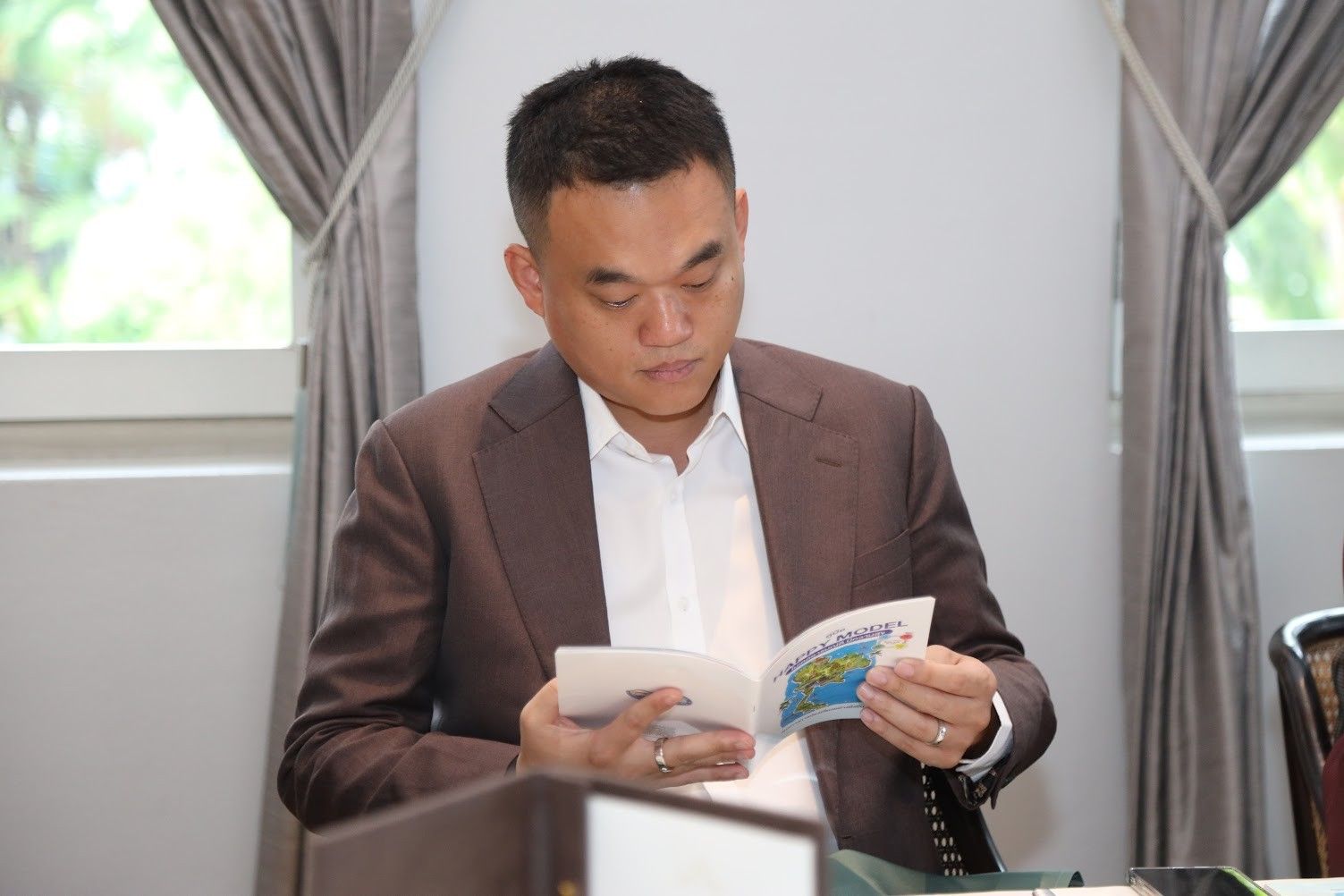 Man in brown suit reading a colorful booklet at a table.