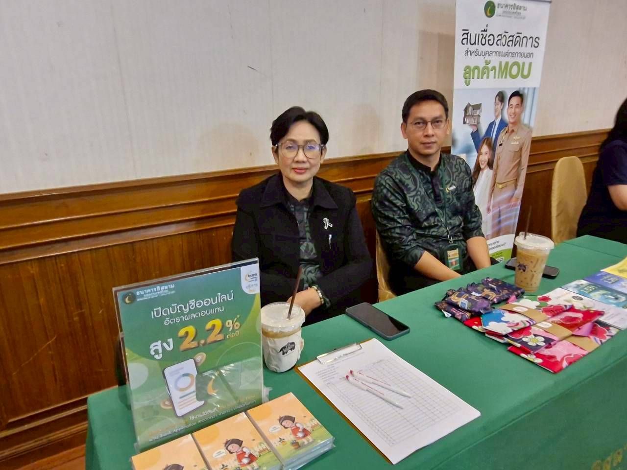 Two representatives sit behind a green table with promotional materials and a background banner.