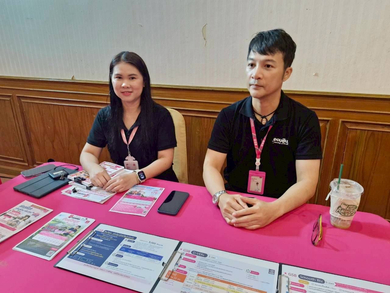 Two people in black shirts sit at a pink table with promotional materials.