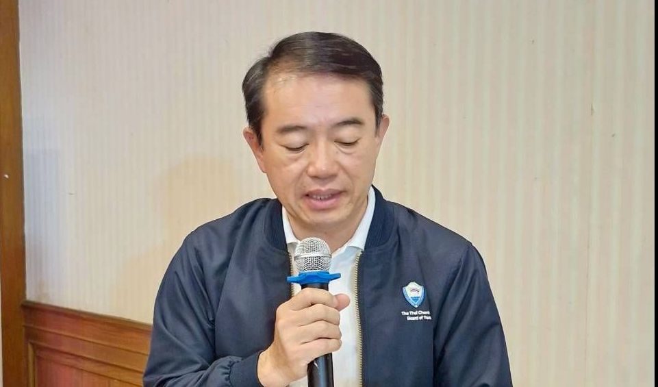 Asian man in dark blue jacket speaking into a microphone while holding documents.